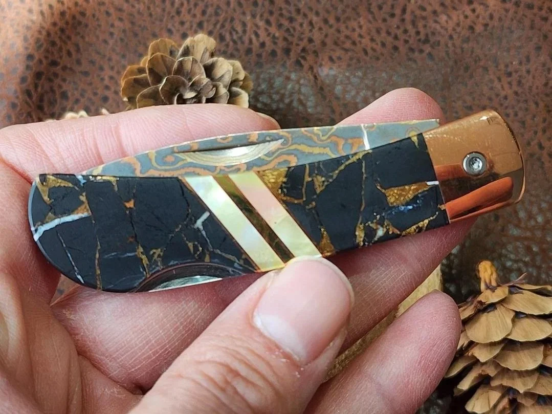 hand holding black buffalon inlay with mother of pearl accents on folding damascus pocketknife with pinecone and wood in back ground