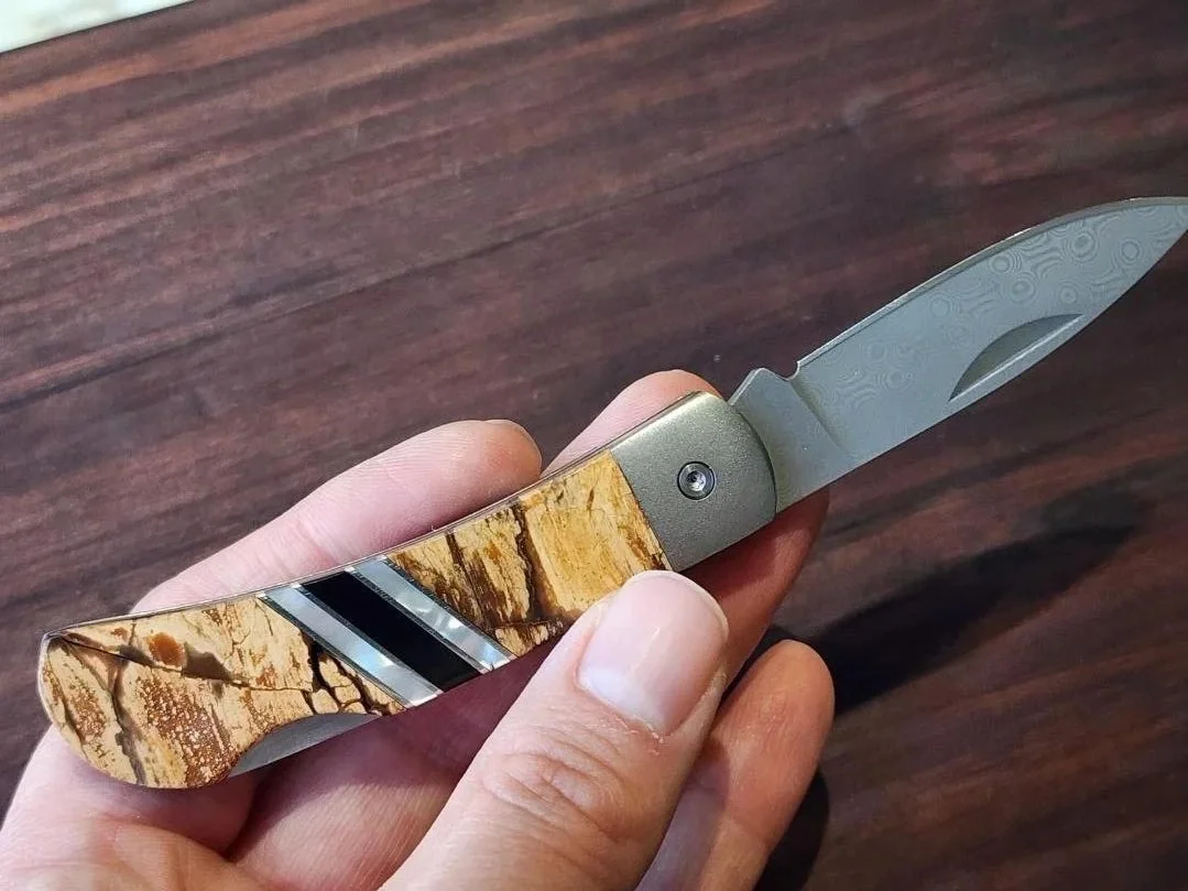 hand holding Damascus folding pocket knife with mammoth handle and raindrop Damascus blade with wooden background