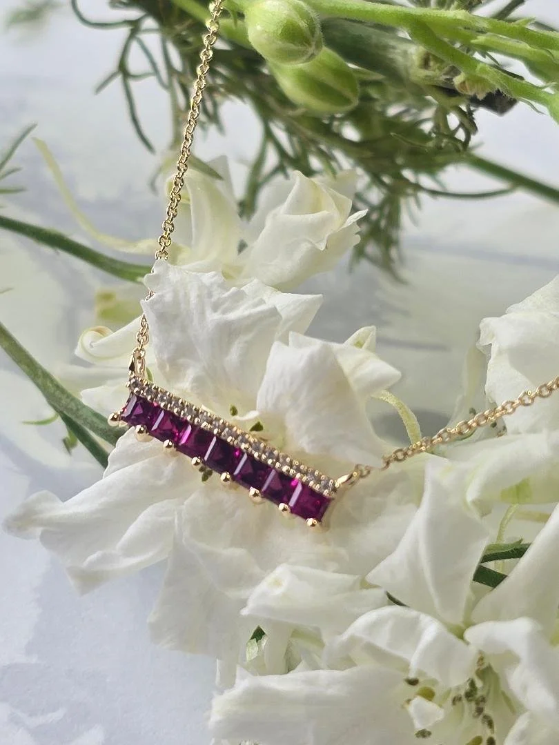 Side view of a yellow gold ruby and diamond bar style pendant draped over white flowers