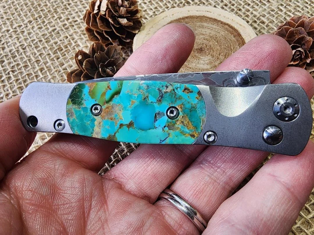hand holding One 3.5" damascus knife with a turquoise inlay on burlap with a wood slice and pinecones in background