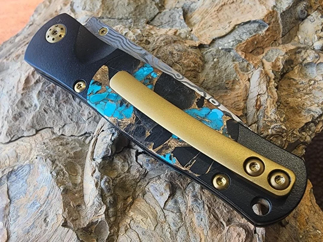 back of black folding knife with Damascus blade with turquoise onyx and copper on the handle and a clip on rock