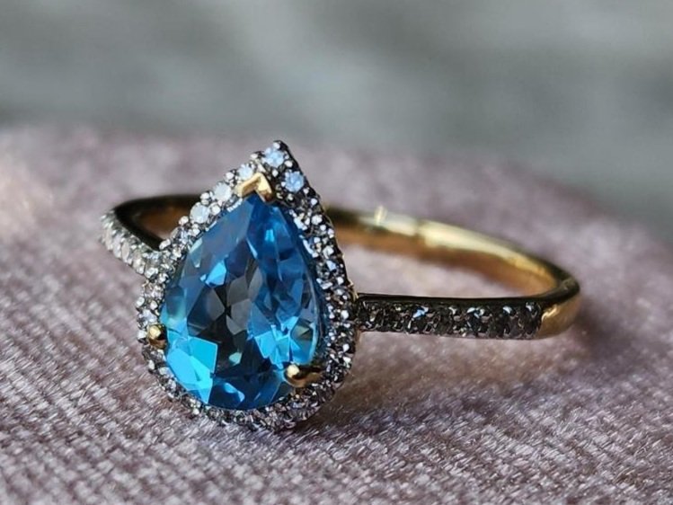 close up yellow gold ring with pear shaped blue topaz and diamond halo and shoulders accents on a pink box