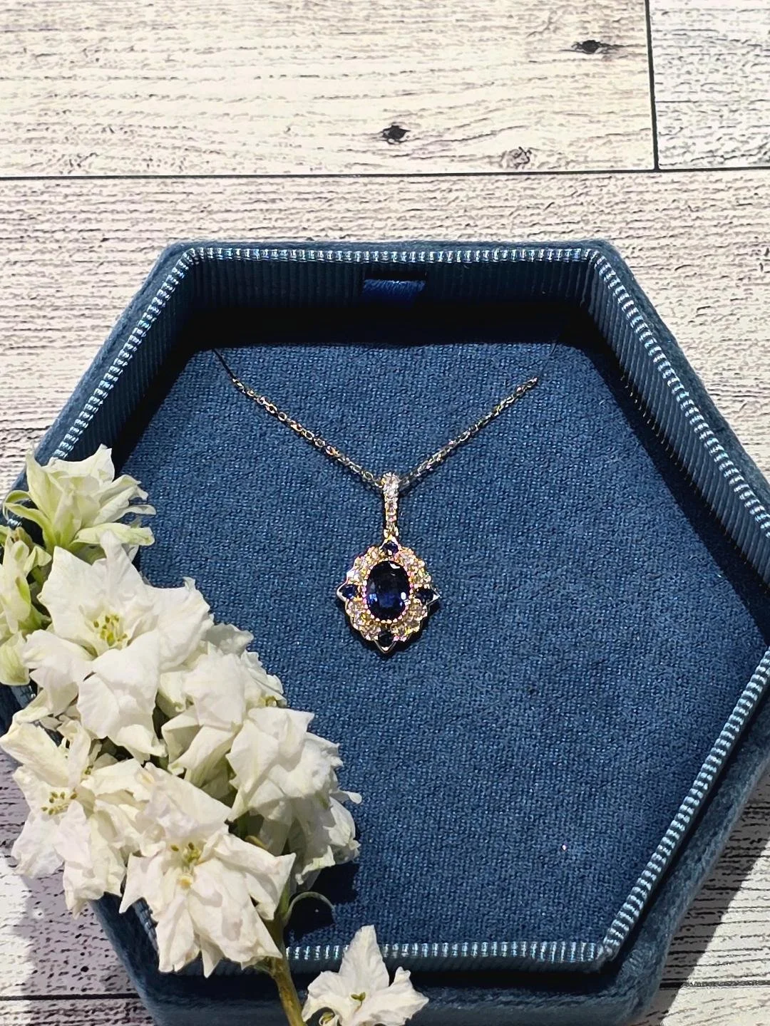 Front view of yellow gold sapphire pendant in a navy blue box with a white wooden back ground