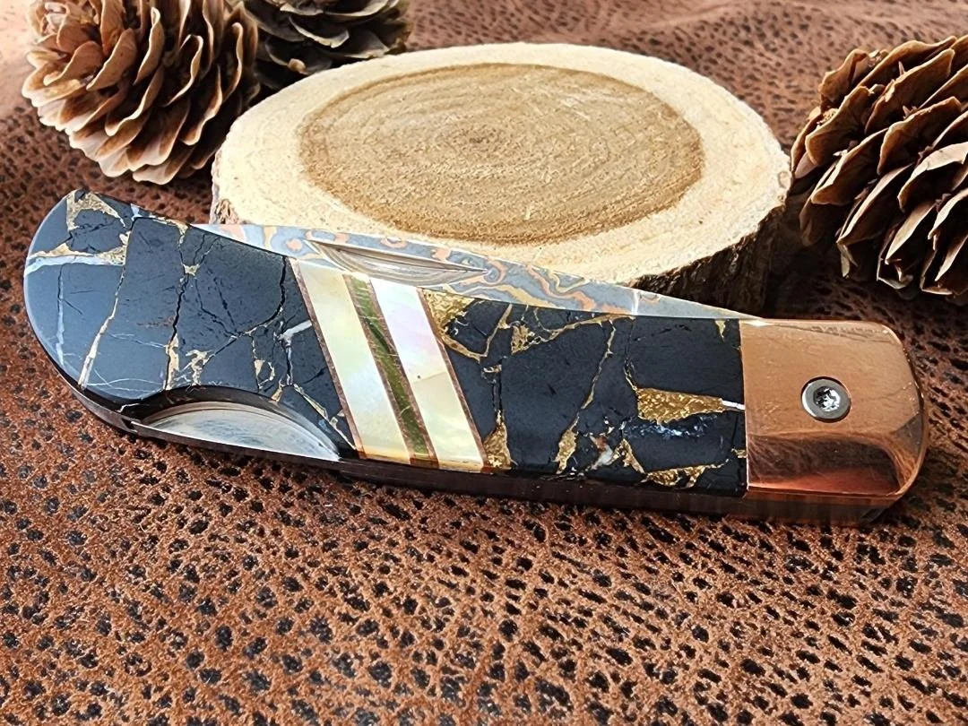 black buffalon inlay with mother of pearl accents on folding damascus pocketknife with pinecone and wood in back ground