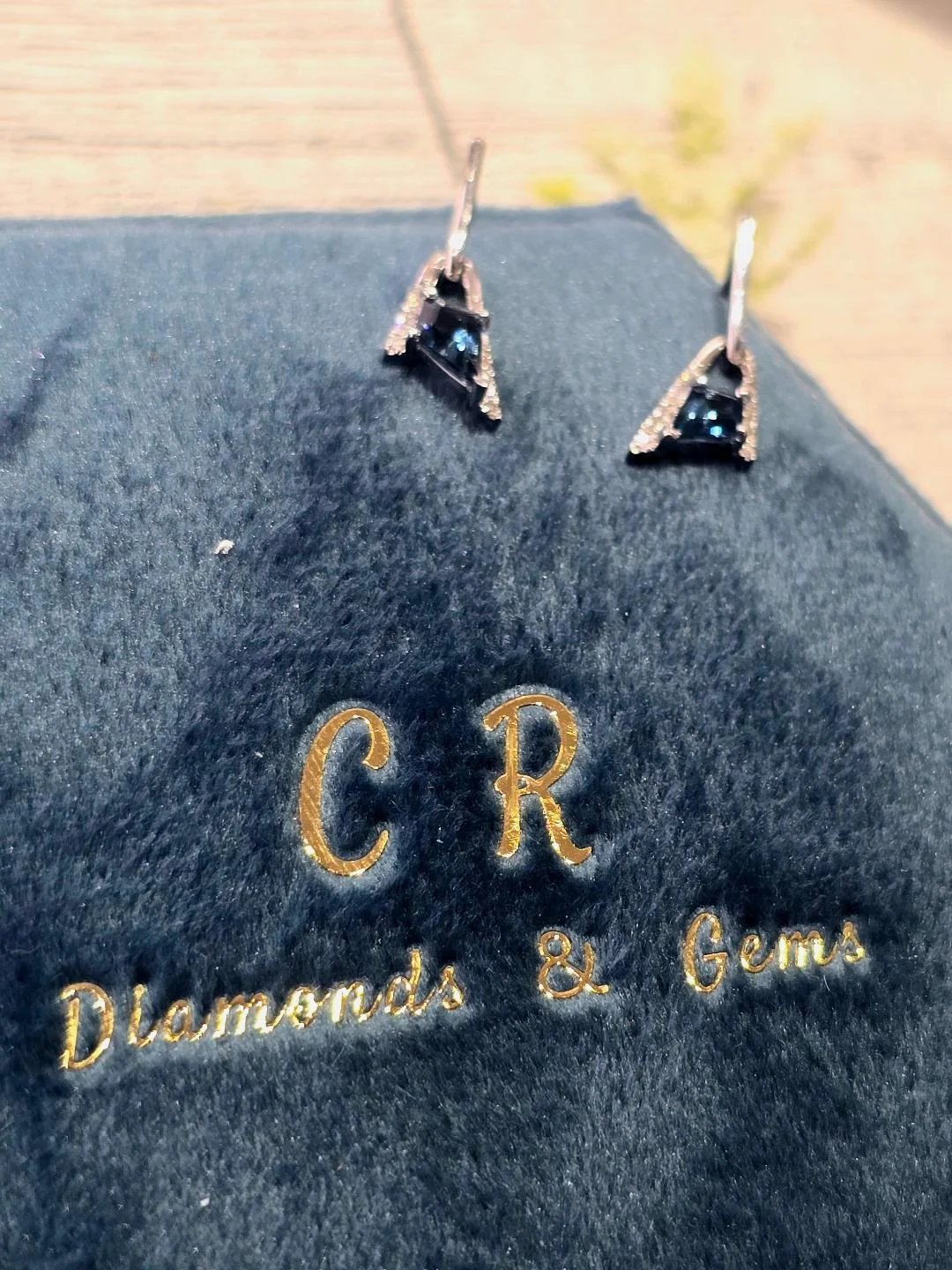 Angled view of white gold London blue topaz earrings in a navy blue box with a white wooden background