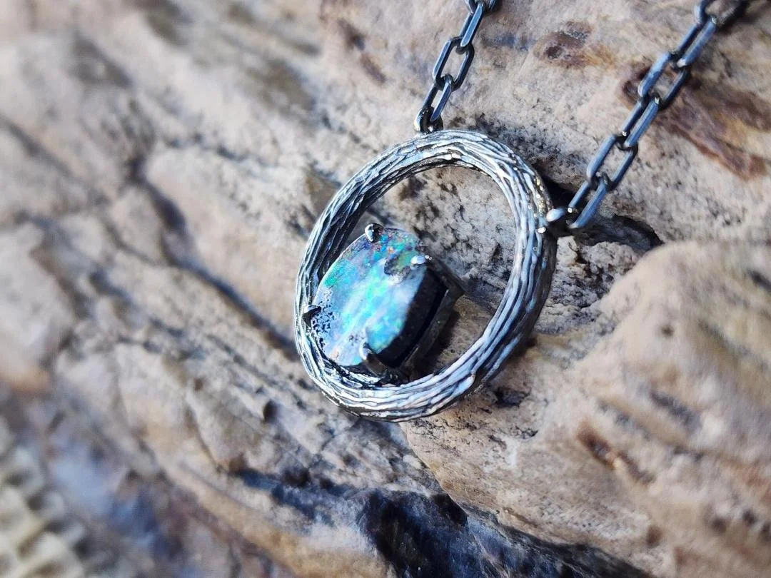side view Blackened sterling silver necklace with a circle pendant featuring a round boulder opal displayed on a rock