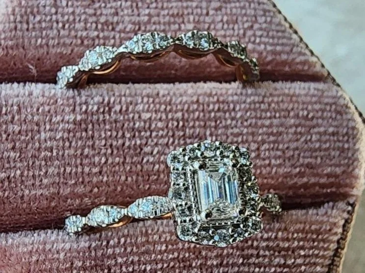 side view White and rose gold diamond wedding ring set with a emerald cut center diamond in a halo of round diamonds in a pink box on a grey background