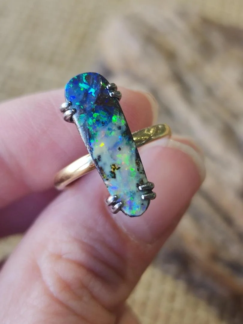 Blue and white elongated Australian boulder opal ring in blackened sterling silver and yellow gold held in between womens fingers with a rock background