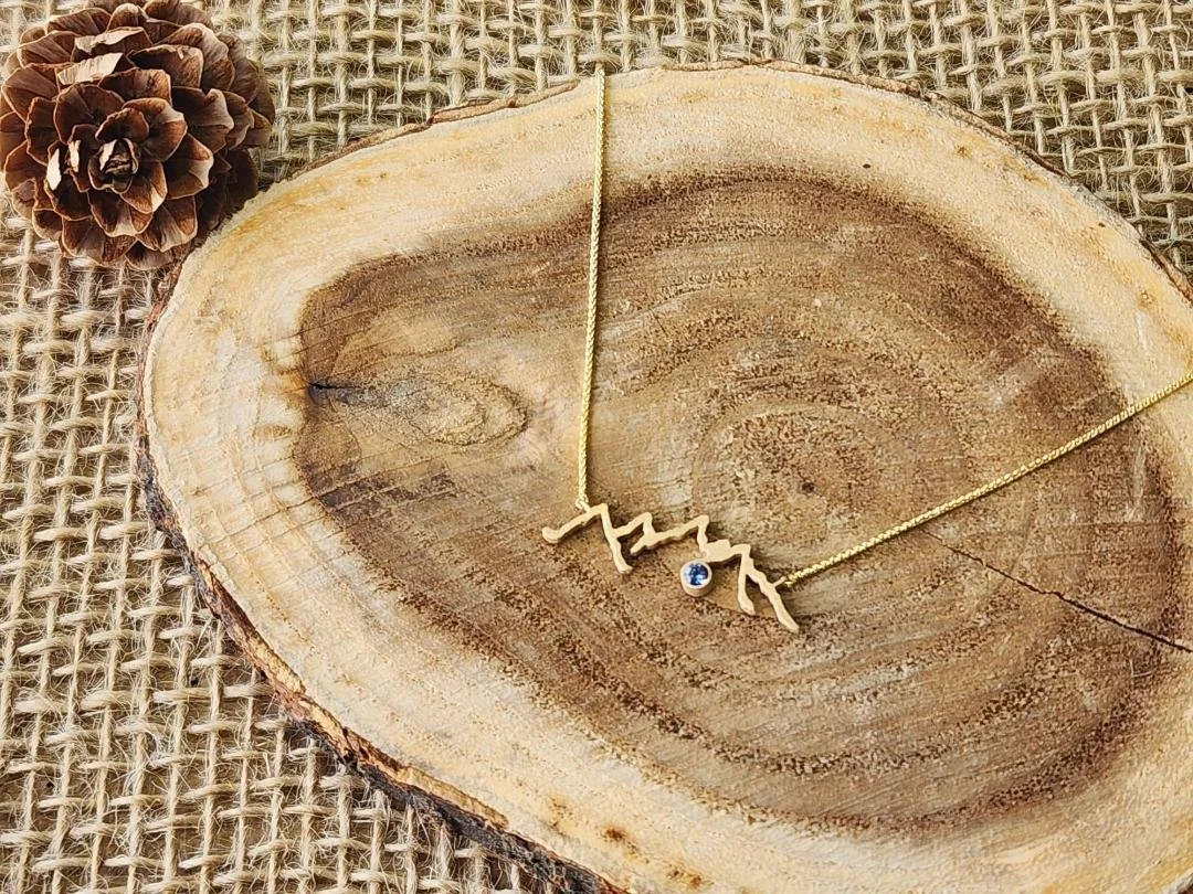 Yellow gold mountain range necklace with round blue sapphire on wood slice and pinecone and burlap in the background