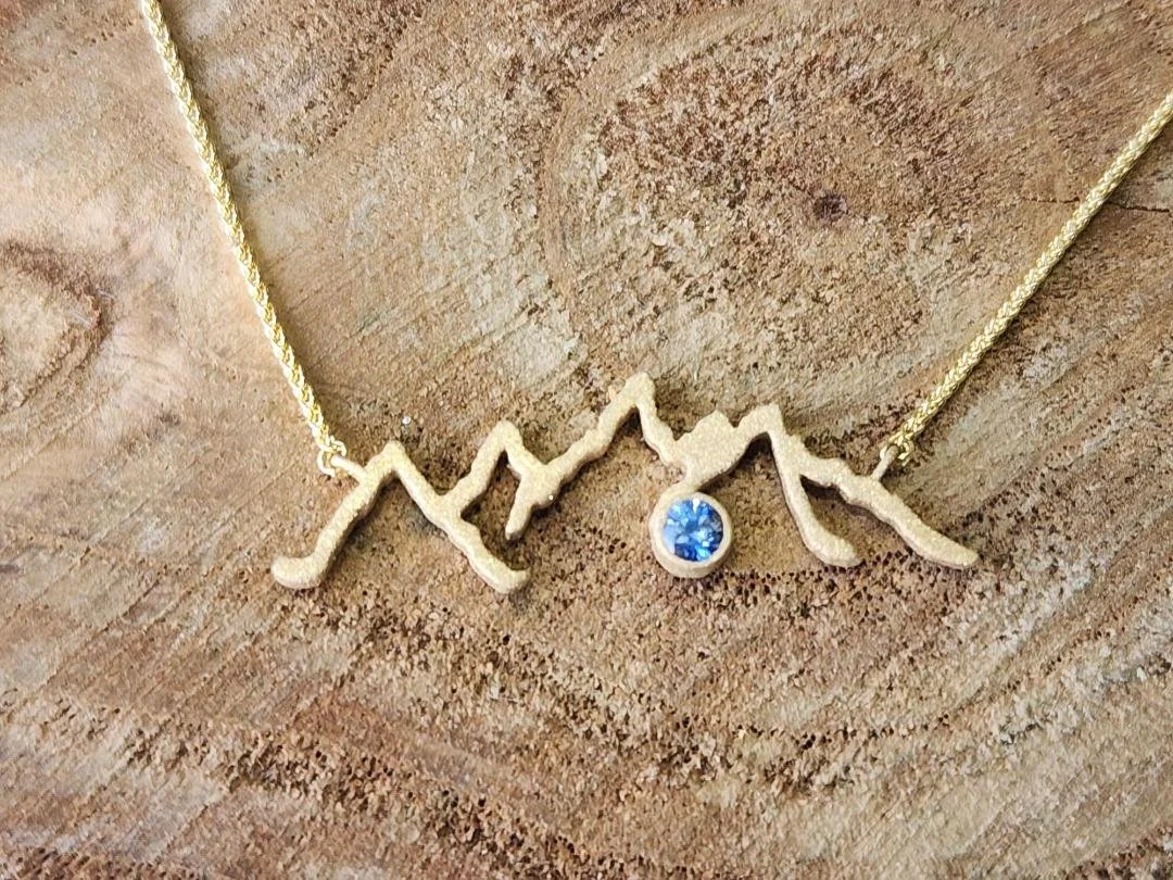 Yellow gold mountain range necklace with round blue sapphire on wood slice