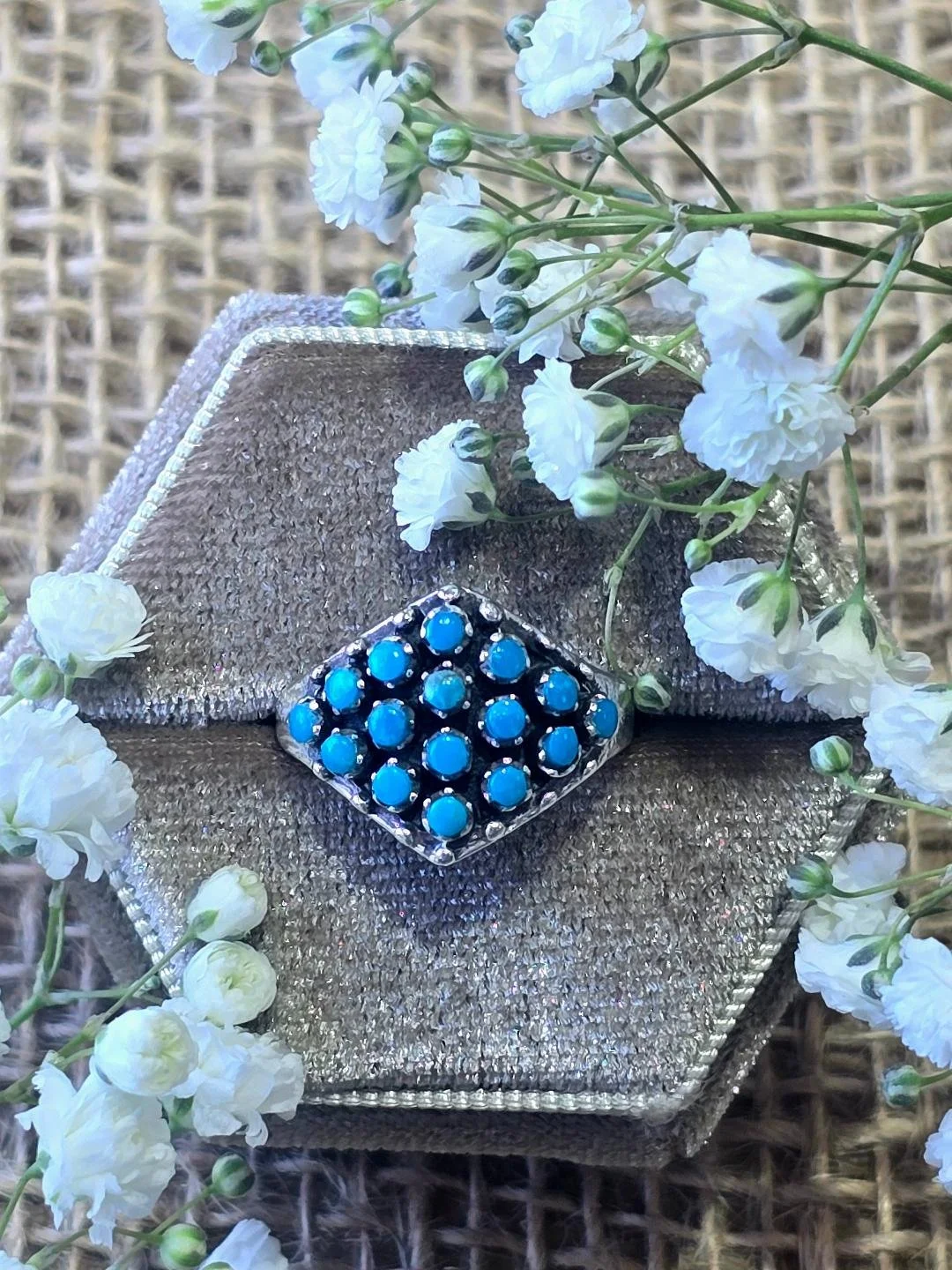 Front view of silver turquoise ring in a brown box with small white flowers set on top of the box
