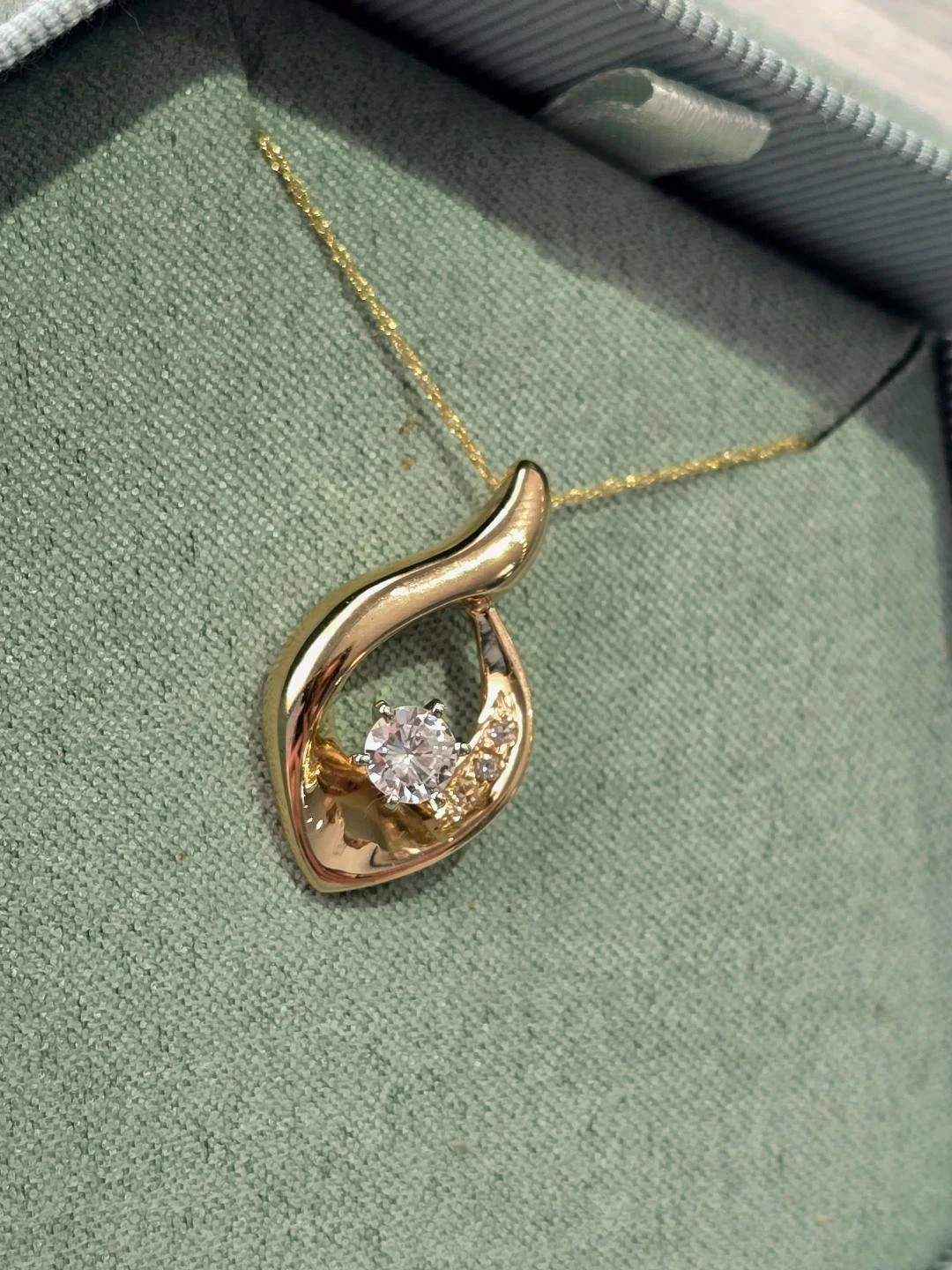 Side view of yellow gold drop pendant in a sage green box with greenery around the box