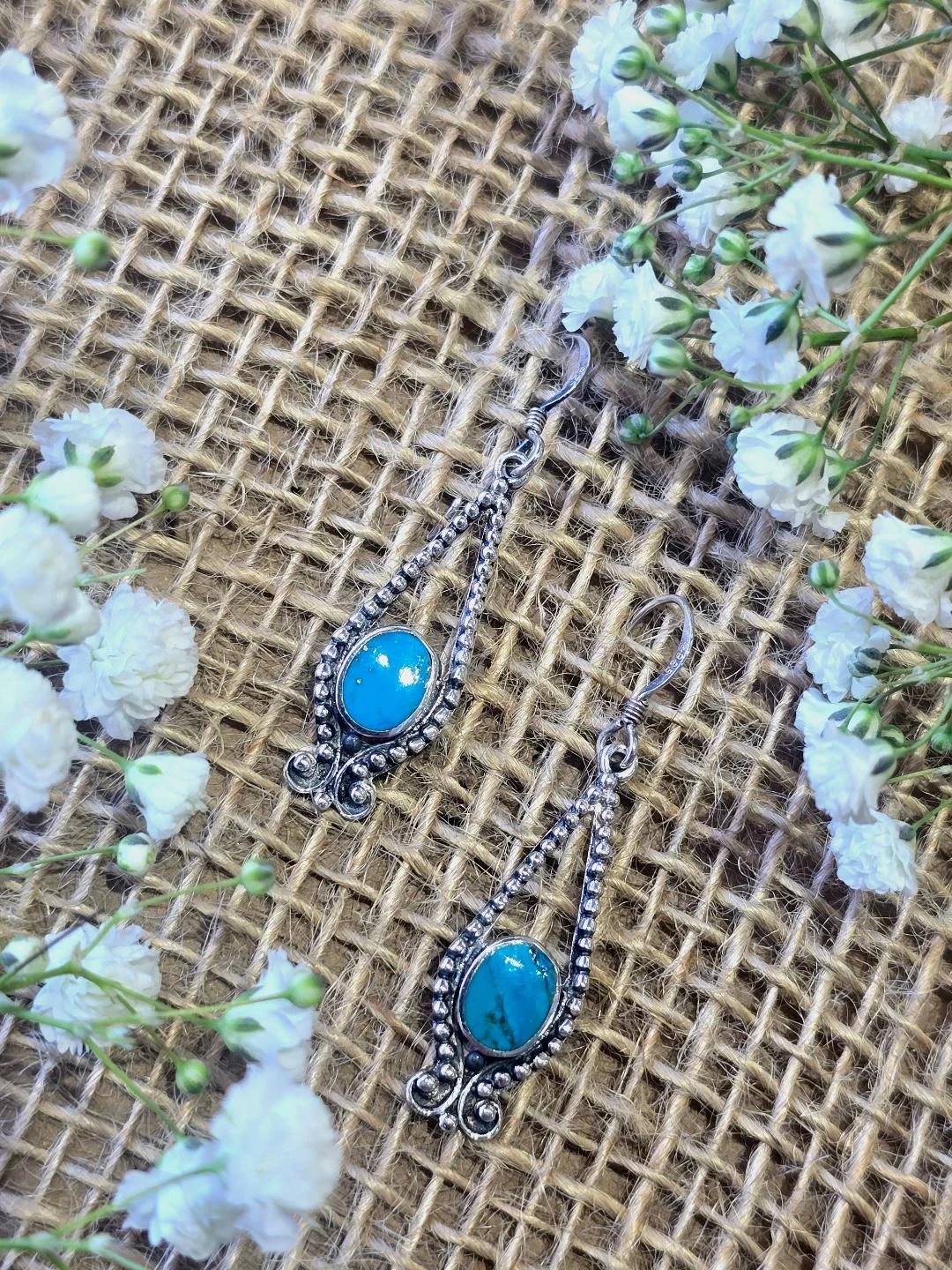 Angled view of silver turquoise dangle earrings on a burlap background with white flowers on the corners