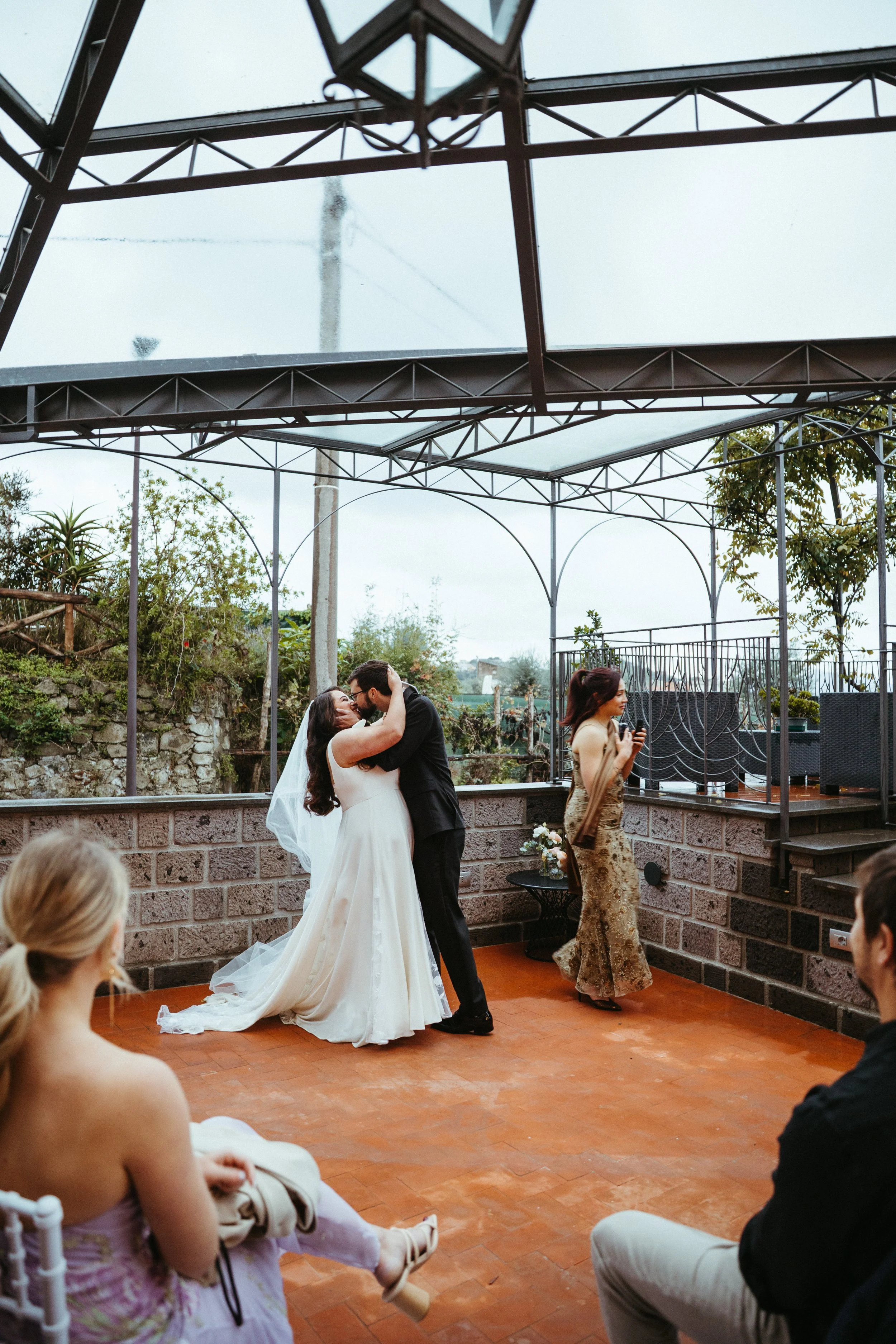 A bride and groom embrace and kiss during their wedding ceremony, with a bridesmaid and guests watching in an outdoor venue.