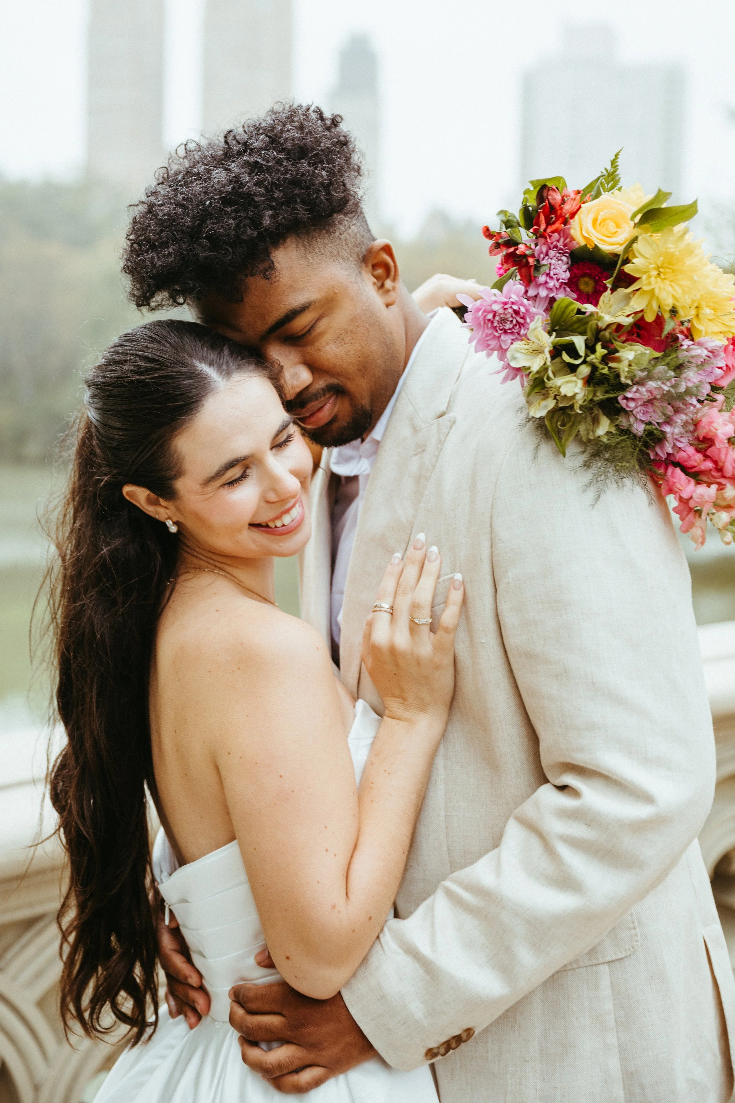 A smiling bride and groom embracing outdoors, with the groom carrying a bouquet of colorful flowers on his shoulder.