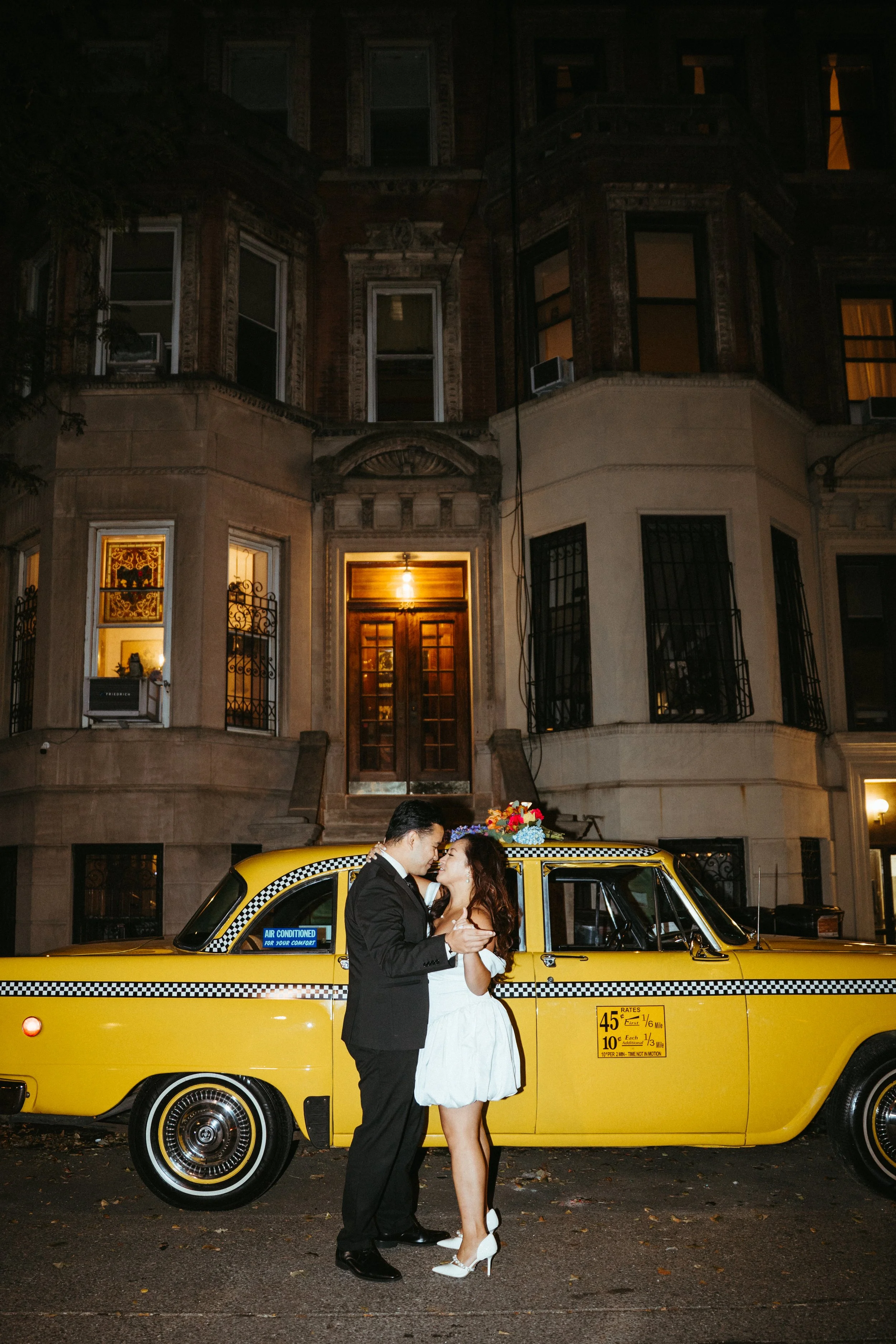 A couple dressed in formal attire, the man in a black suit and the woman in a white dress, are dancing and smiling in front of a yellow taxi cab with a bouquet of flowers on top. The setting is at night in front of a residential building with lit win