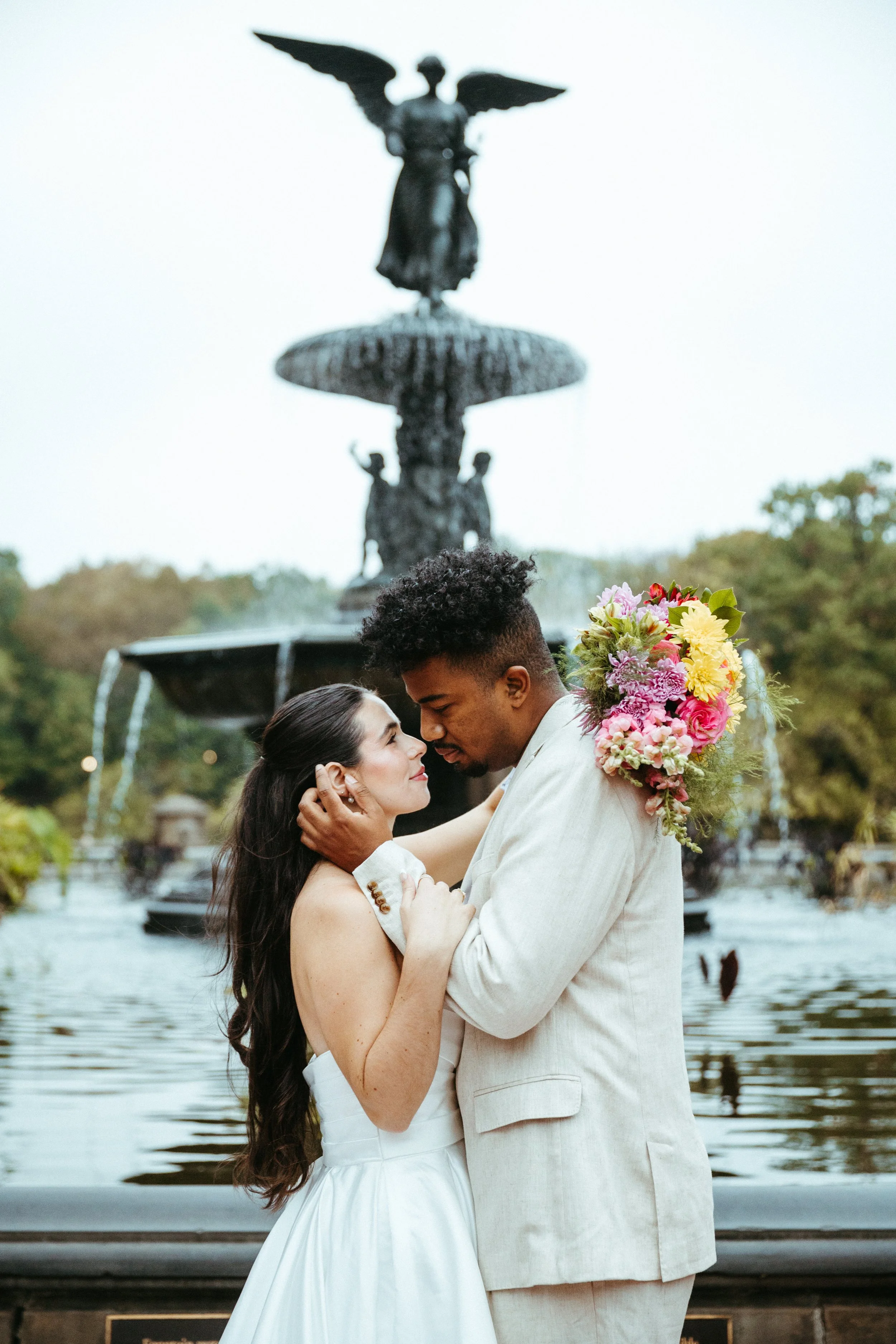 A couple embraces in front of a fountain, with the woman holding a colorful bouquet of flowers, during a wedding or romantic photoshoot.