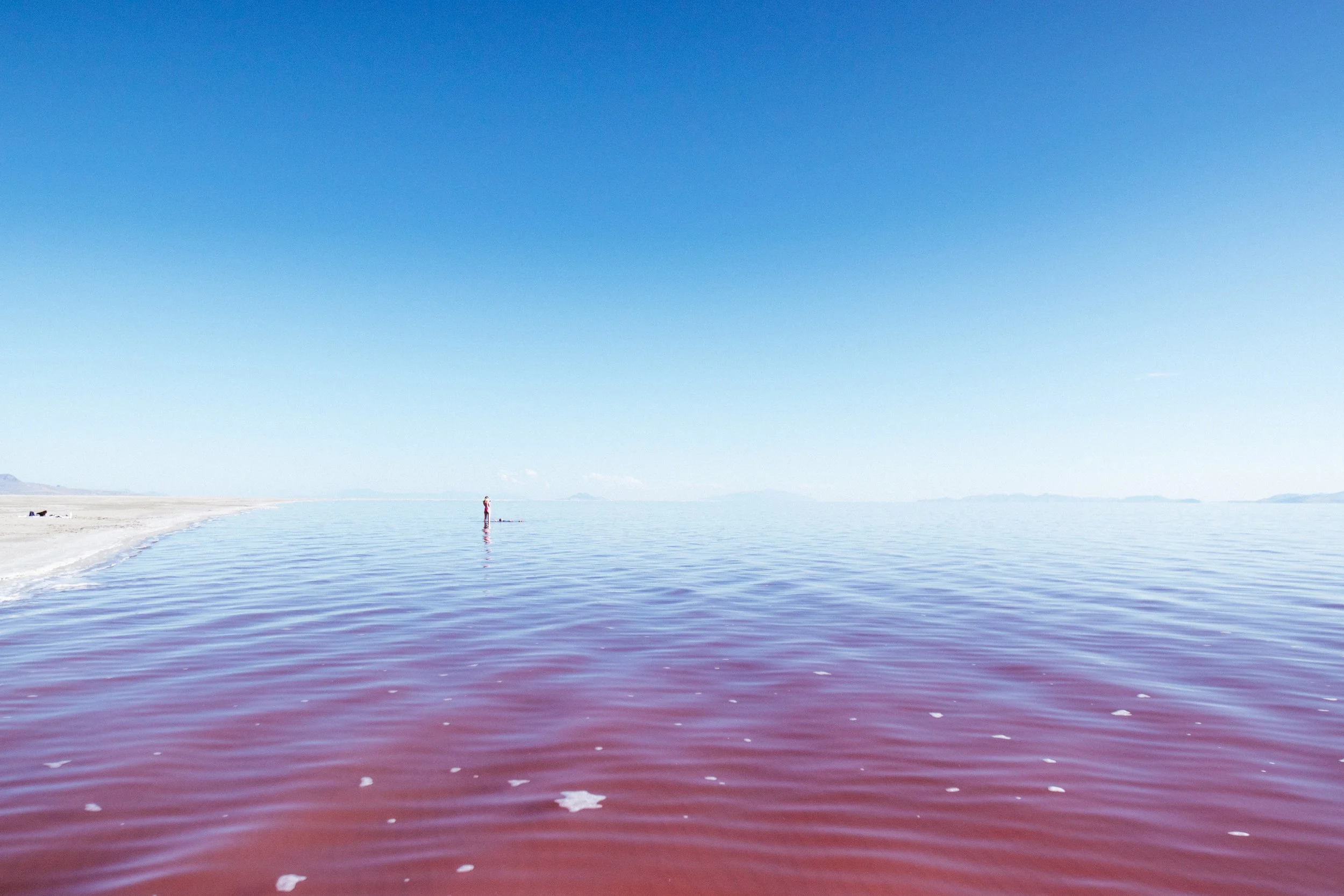 A person standing in shallow water near the sandy shoreline of a salt lake, with pinkish water and a clear blue sky.