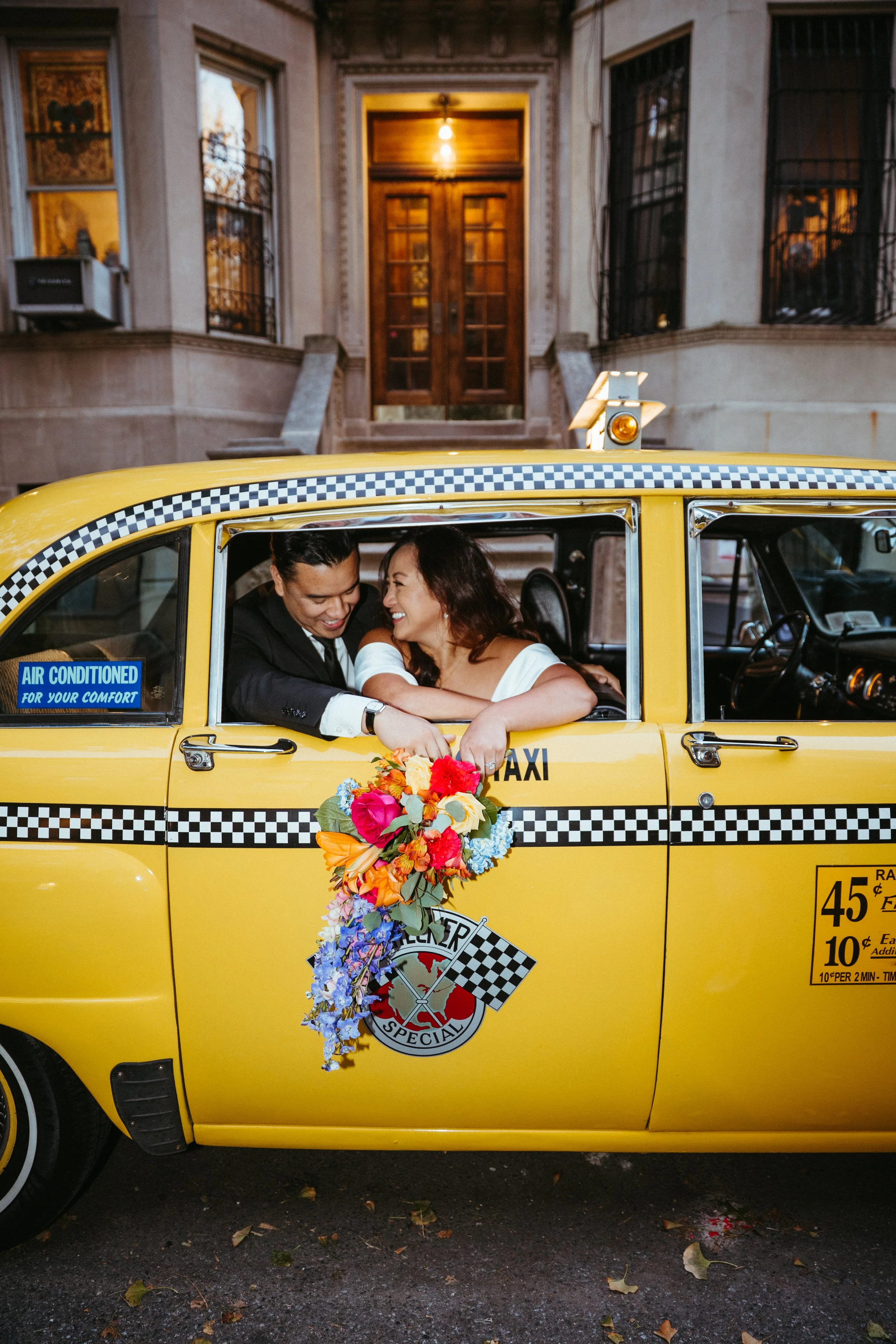 A smiling couple inside a yellow taxi, with the woman holding a colorful bouquet, on a city street in front of an ornate building.