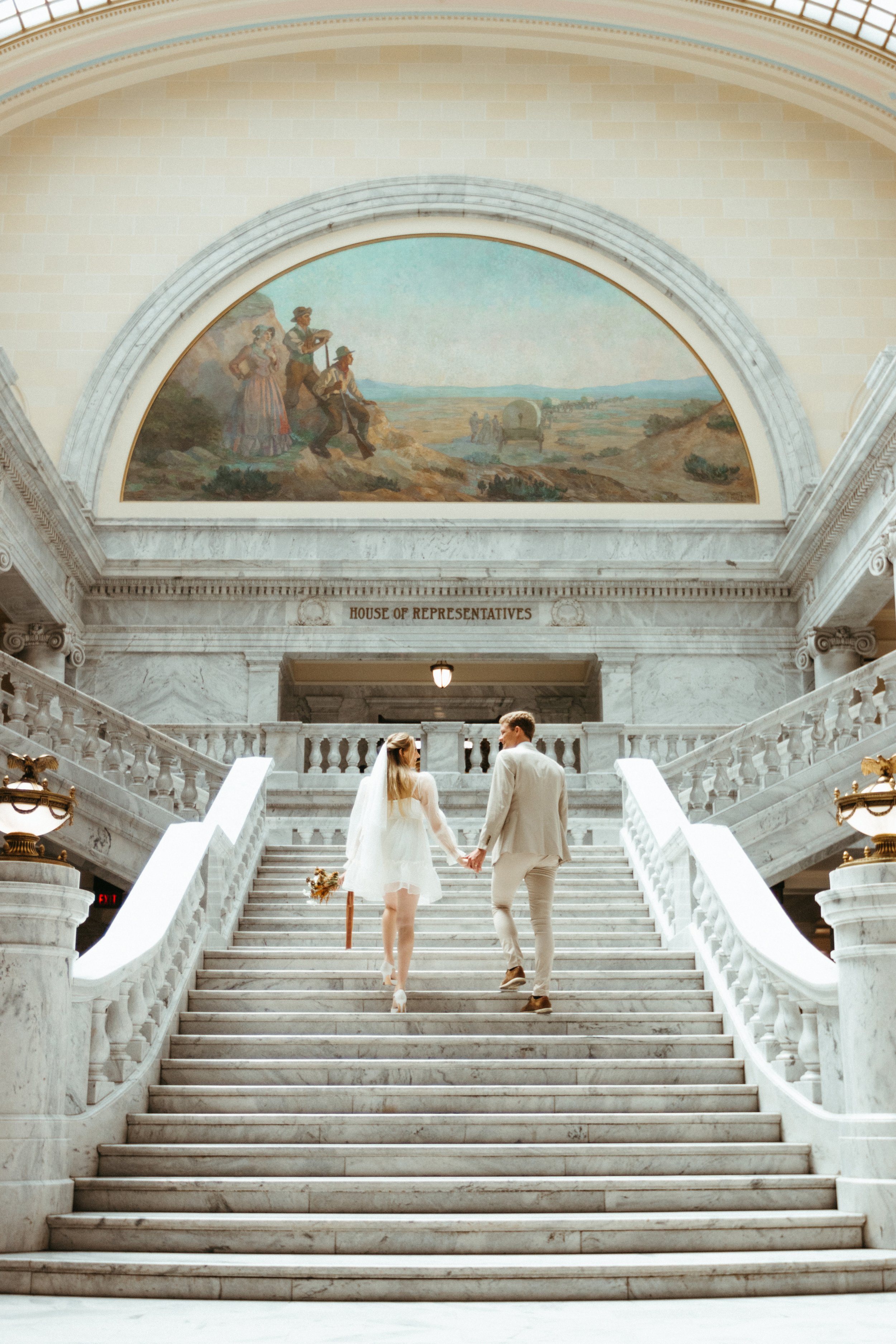 A couple walks hand in hand up a grand marble staircase inside a historic government building labeled 'House of Representatives.' The woman is dressed in a white dress with a veil, and the man is in a light suit. The background features a large mural