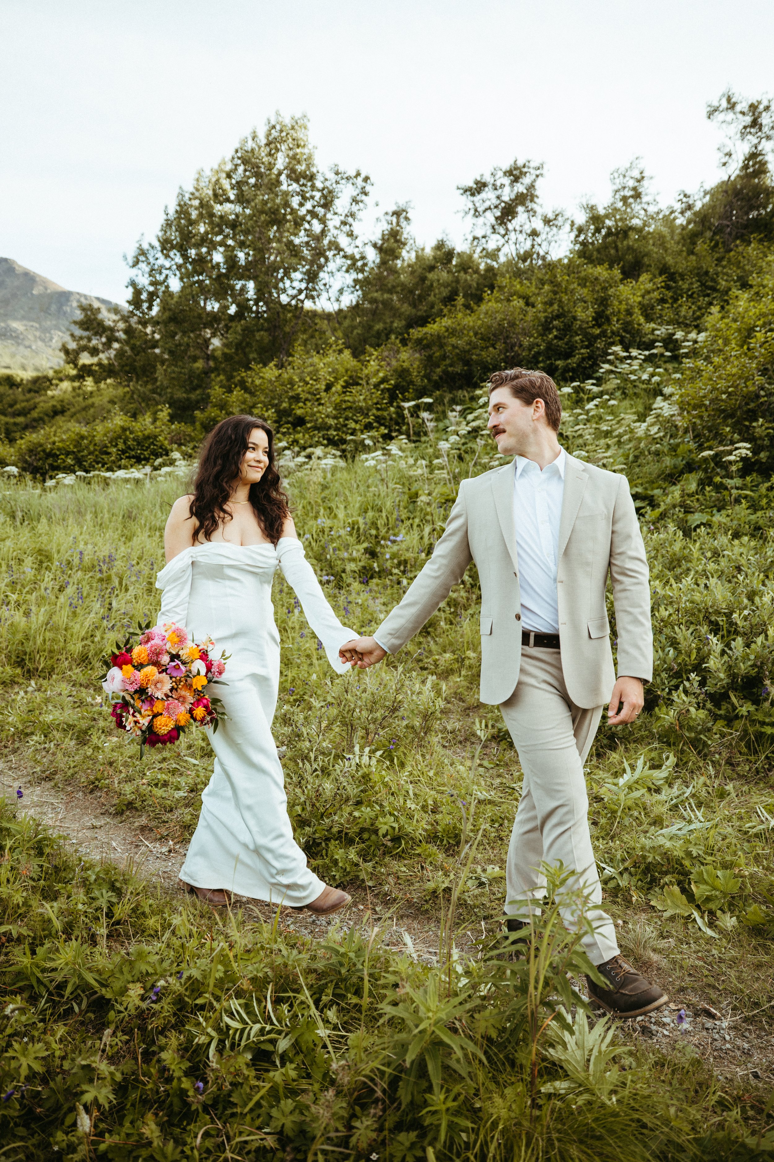 A bride and groom holding hands while walking through a grassy outdoor area with trees and mountains in the background. The bride wears a white dress and holds a colorful bouquet, while the groom wears a light-colored suit.