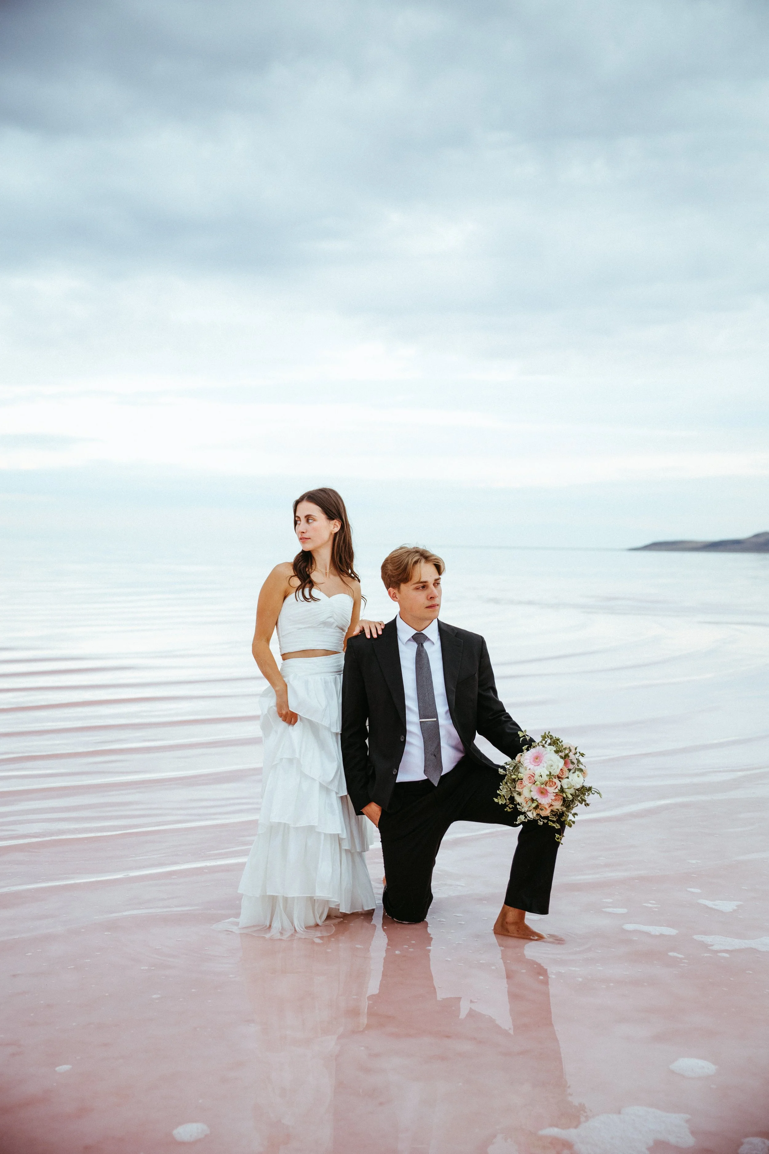 A bride in a white wedding dress and a groom in a black suit are at the beach, with one kneeling and holding a bouquet of flowers, while the other stands beside him.