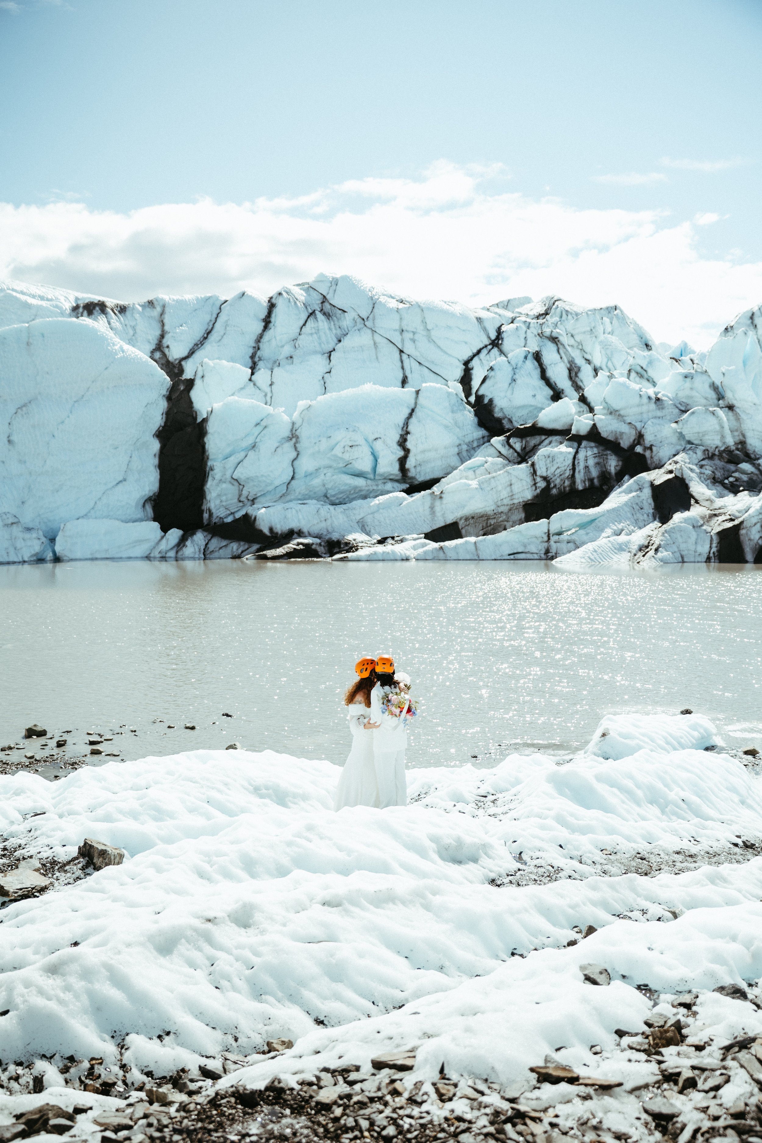 Two women in white dresses and orange helmets holding a bouquet of flowers, standing on snow near a partially frozen lake with glacier and ice formations in the background.