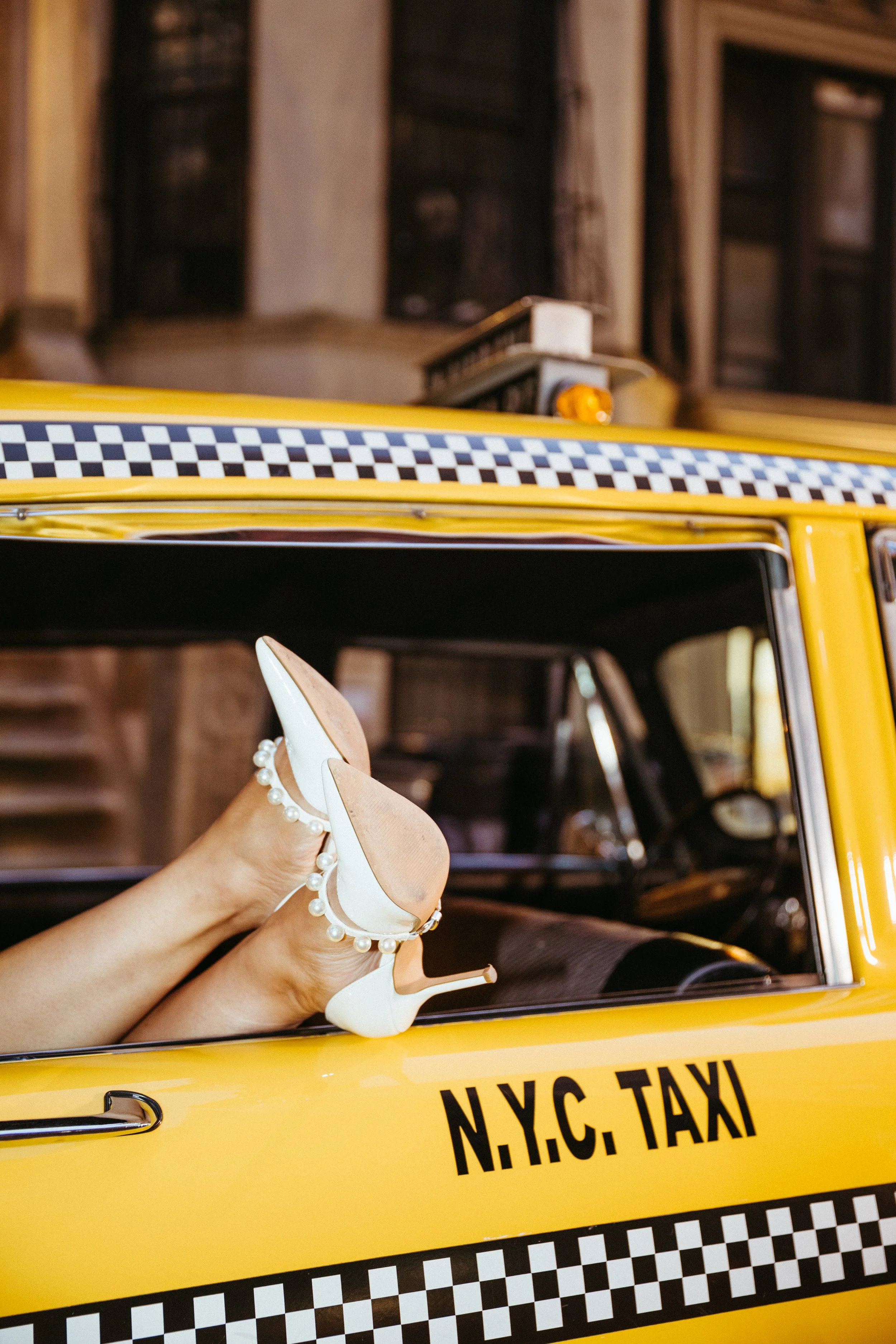 A close-up of a yellow NYC taxi with women's legs and white high heels with pearl accents resting on the window ledge inside the taxi.