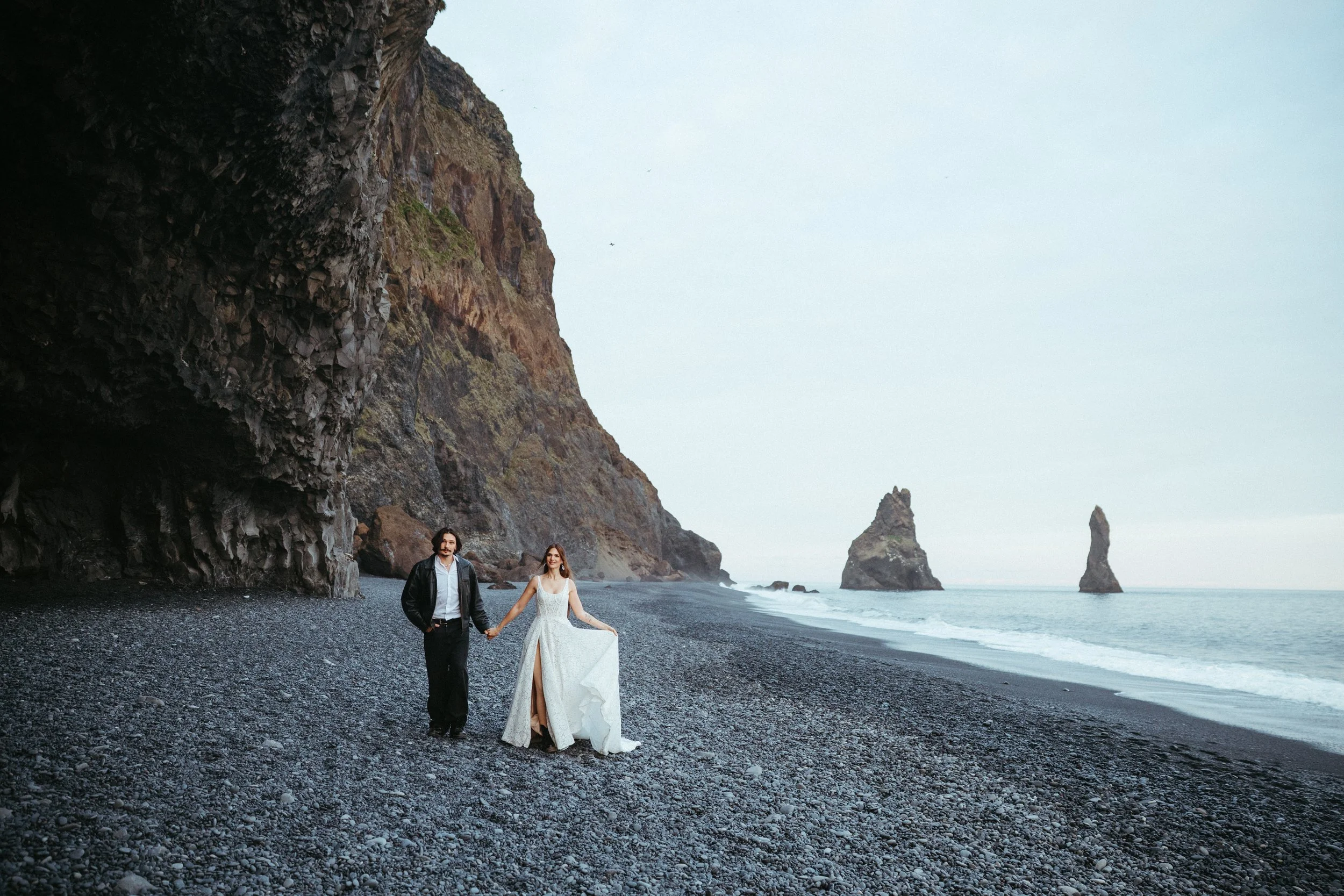 A bride and groom holding hands on a black pebble beach with large cliffside formations and sea stacks in the background.