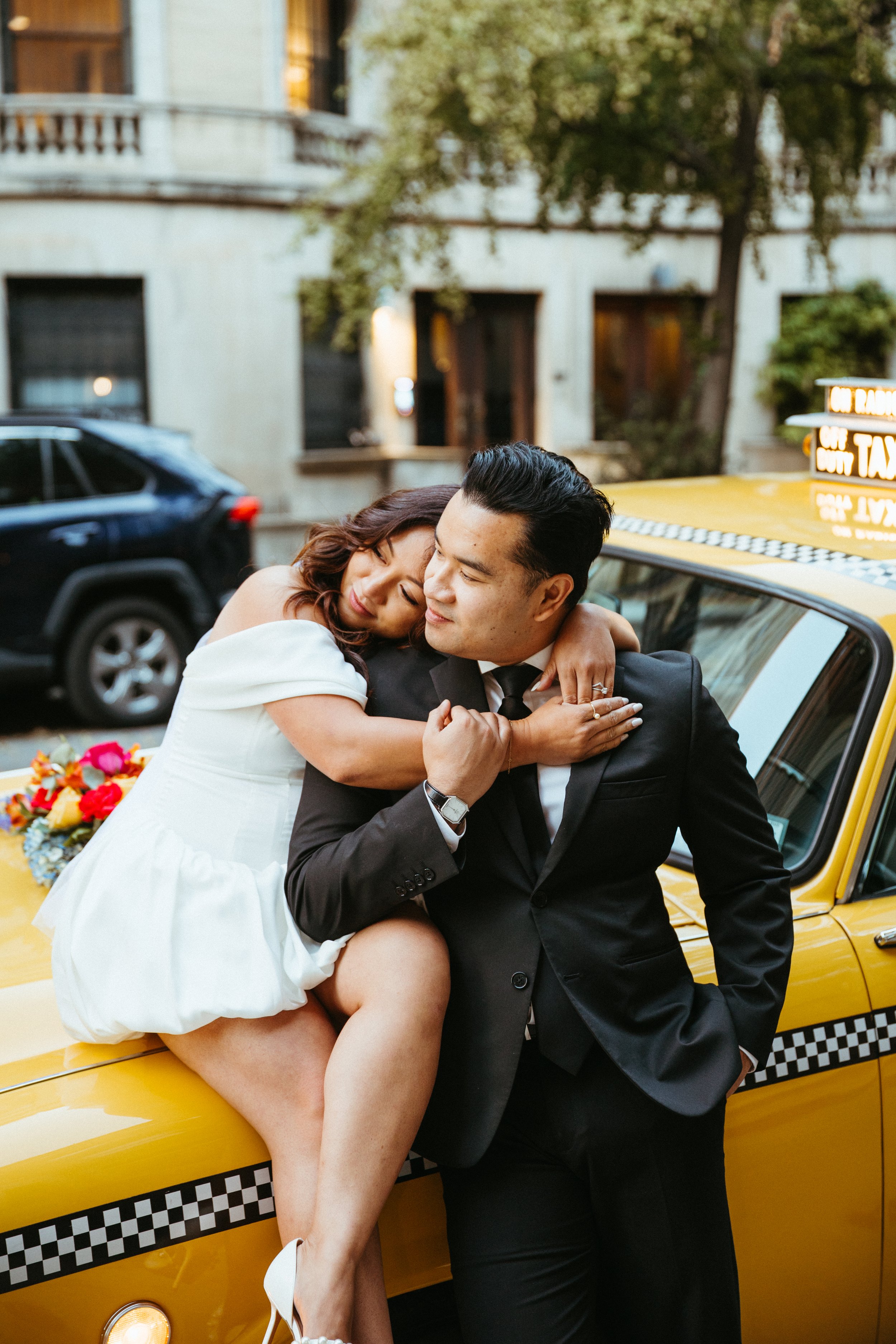 A woman in a white dress hugging a man in a black suit and tie while sitting on a yellow taxi cab.