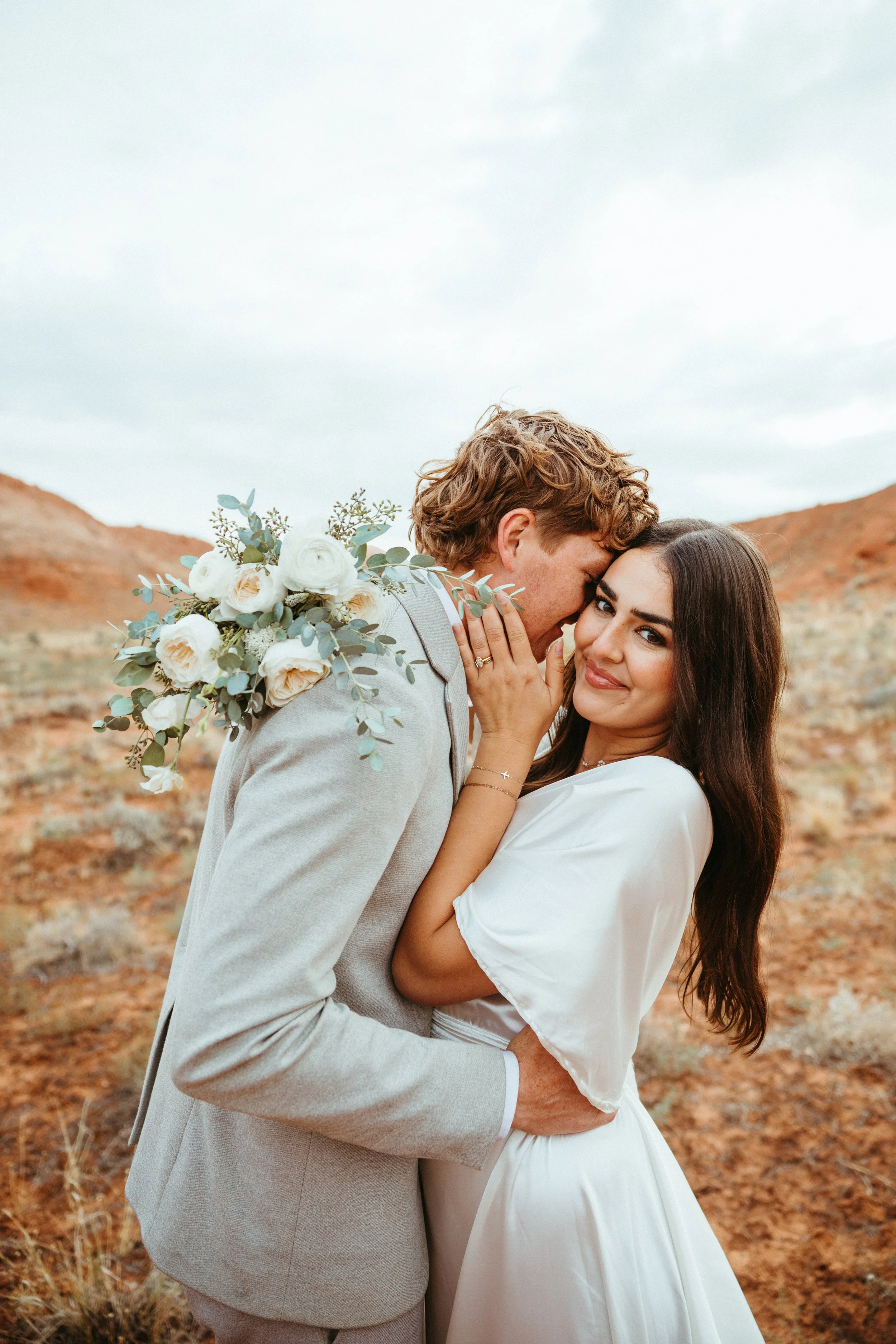 A newlywed couple embracing outdoors, with the groom whispering into the bride's ear, the bride smiling, and the groom holding a bouquet of white roses and greenery, set against a landscape with reddish hills and a cloudy sky.