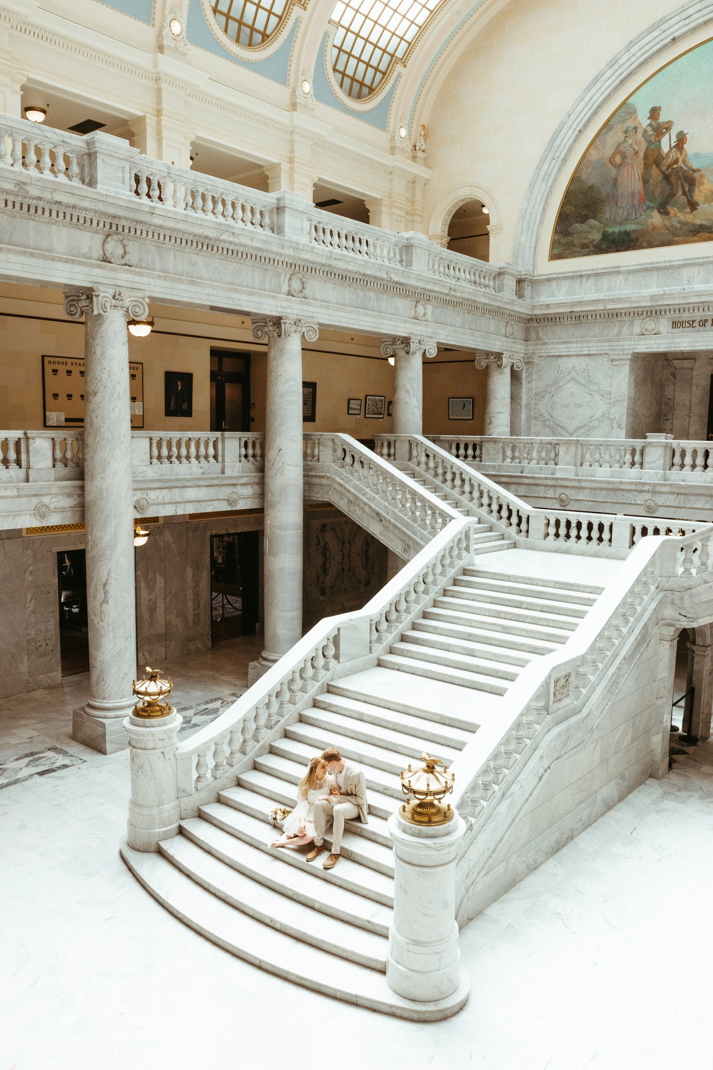 A bride and groom sitting on the marble staircase inside a grand, historic building with ornate columns and a large painting on the wall.