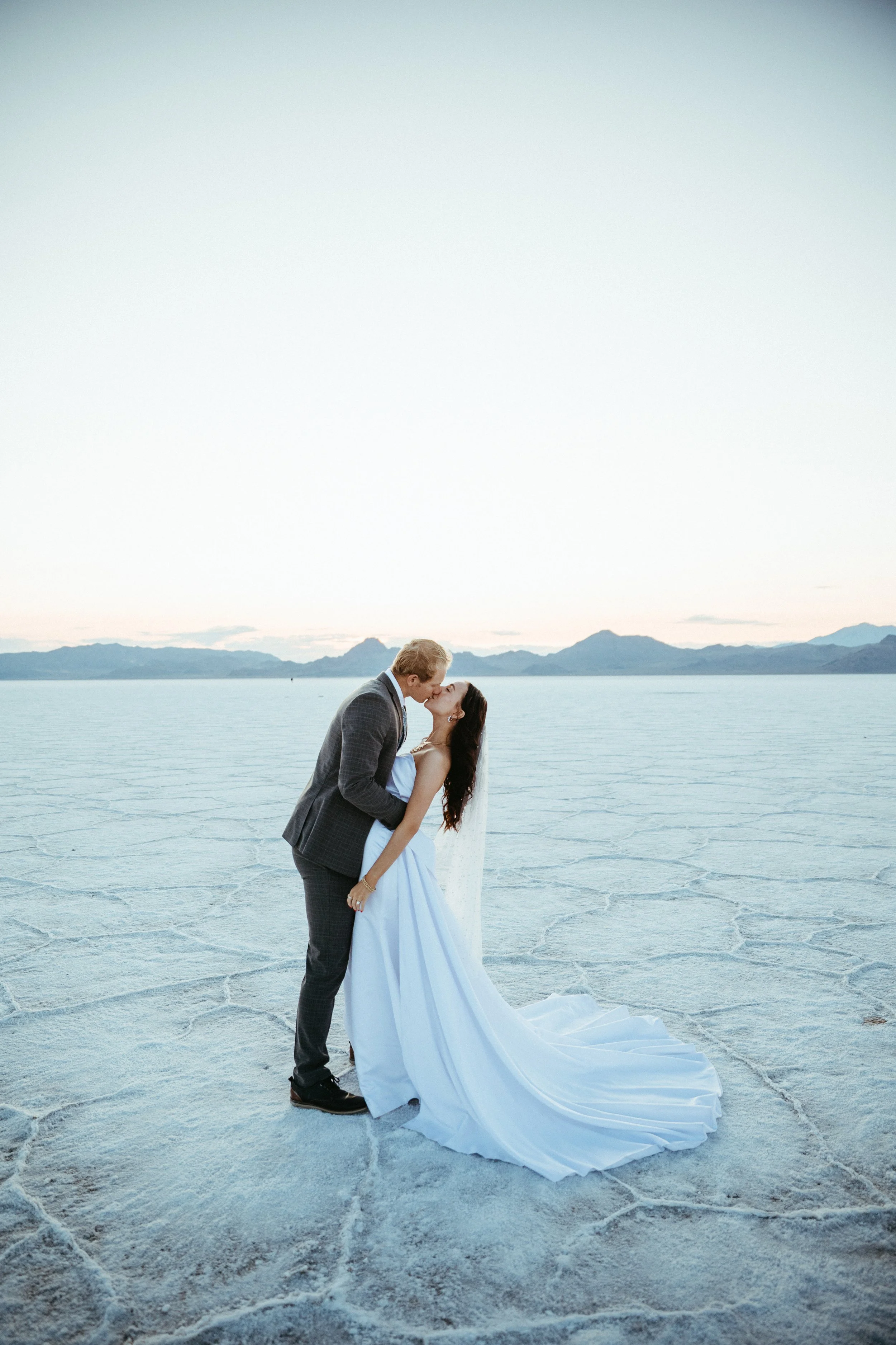 A couple in wedding attire sharing a kiss, standing on a vast salt flat with mountains in the distance during sunset.