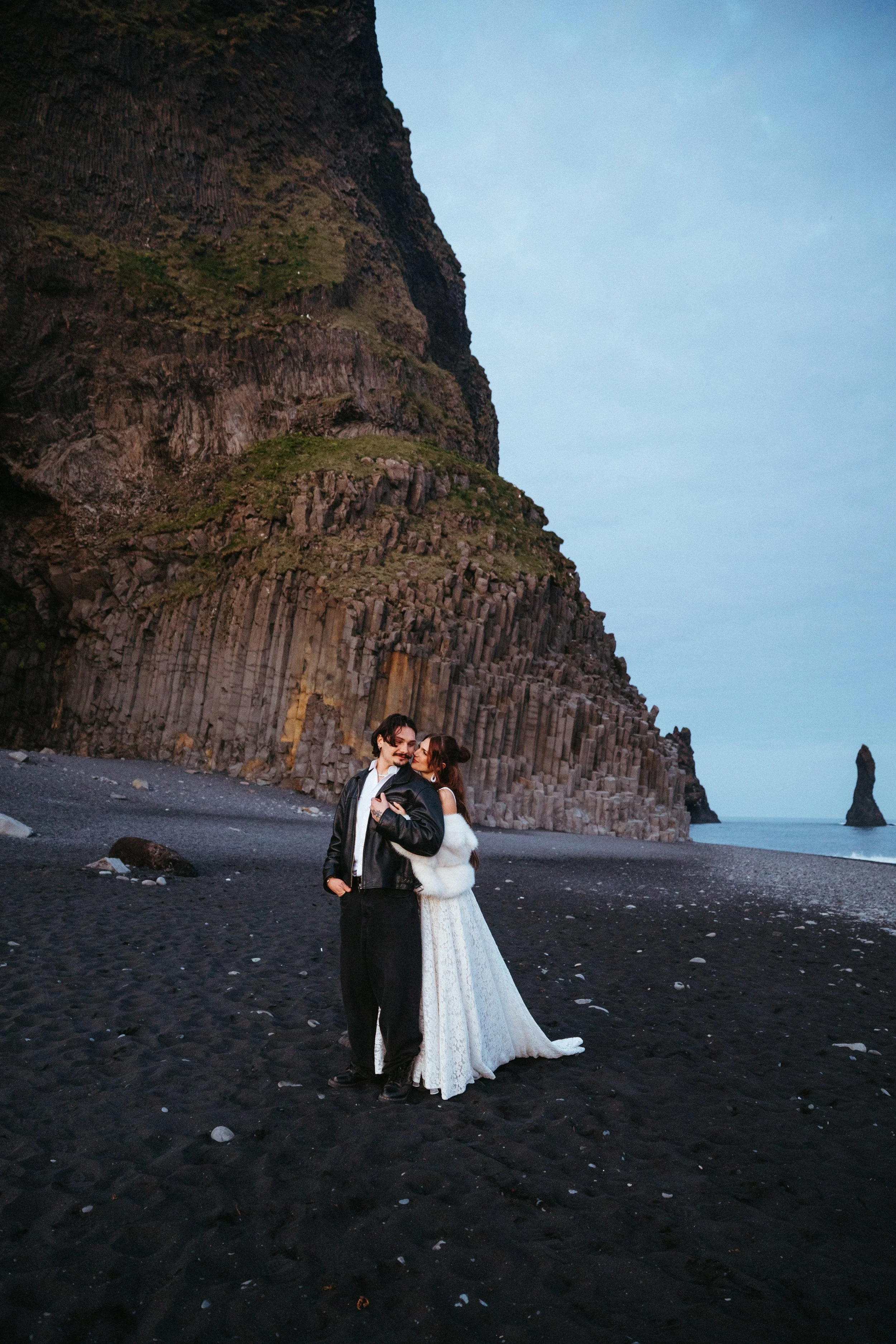 A couple dressed in wedding attire standing on a black sand beach, with a tall rocky cliff and a sea stack in the background