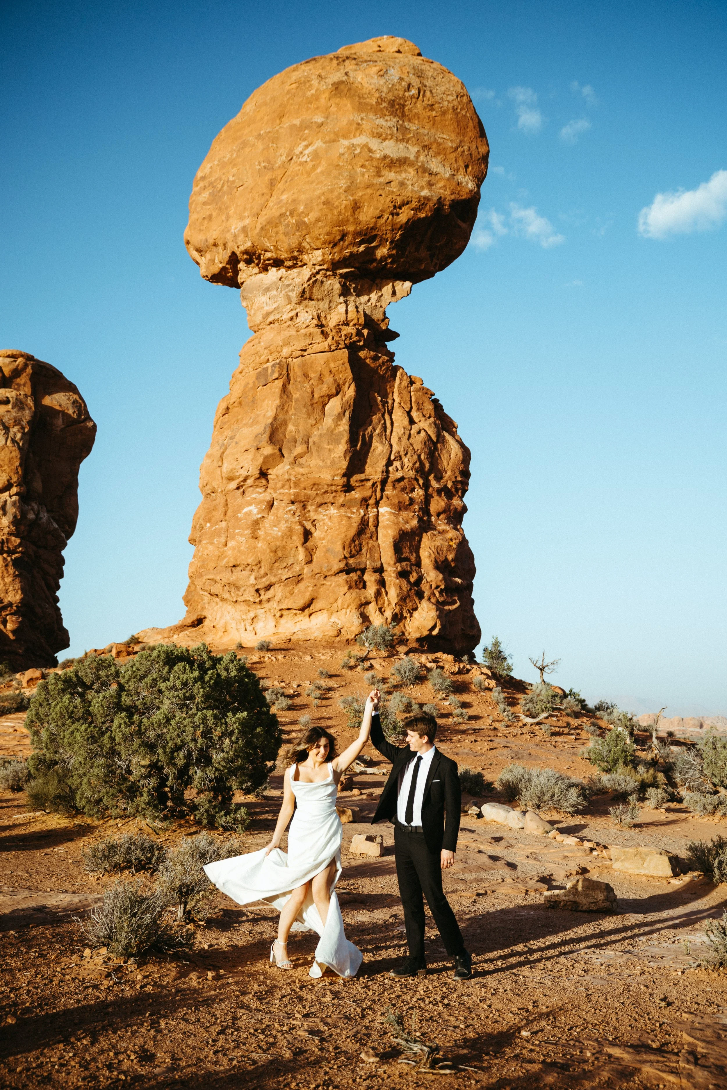 A couple dressed in wedding attire dancing in a desert landscape with large red rock formations and sparse desert vegetation.