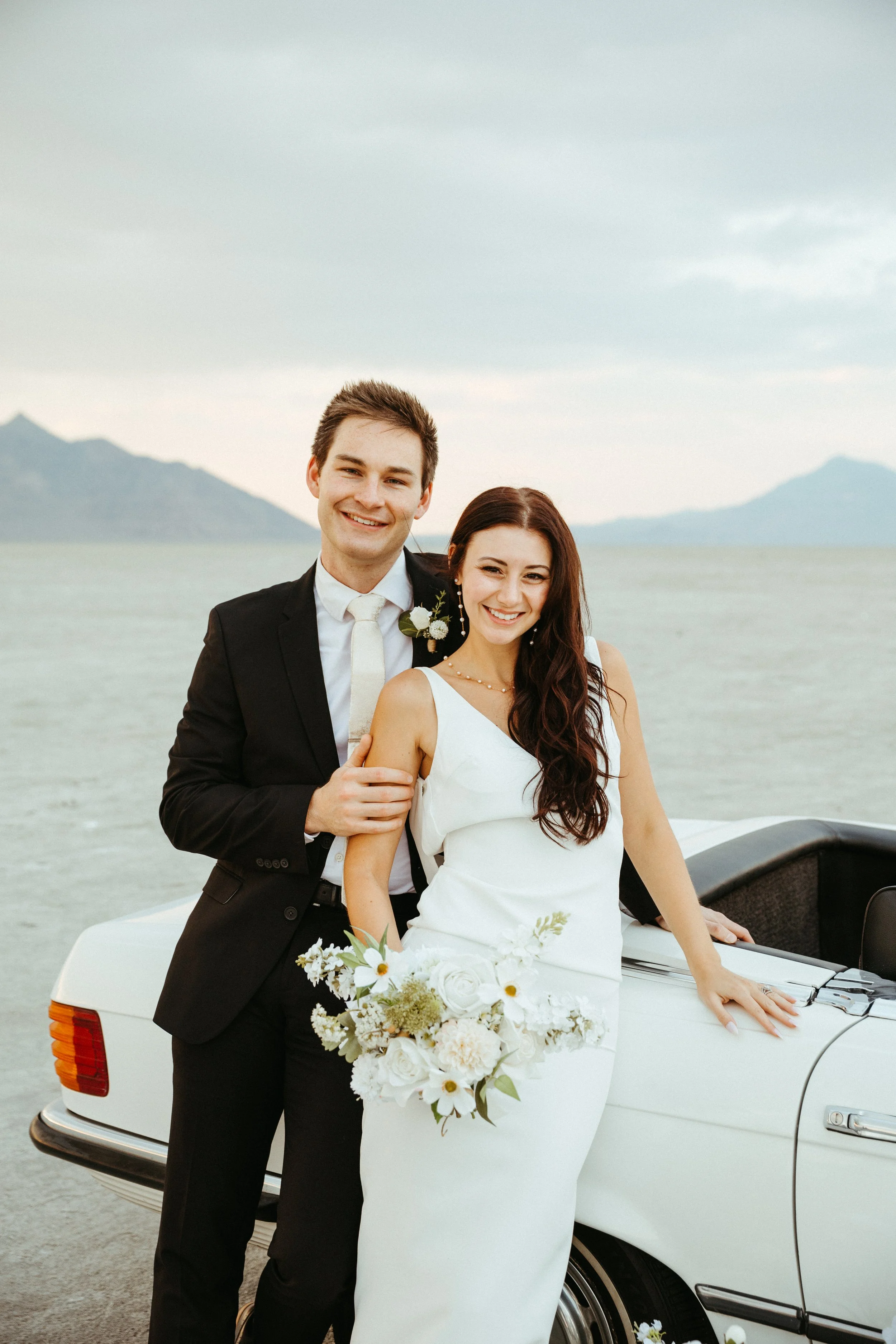 A newlywed couple standing next to a white vintage car near a salt flat with mountains in the background. The groom is in a black tuxedo with a white shirt and tie, smiling, with his arm around the bride. The bride is in a white wedding dress, holdin