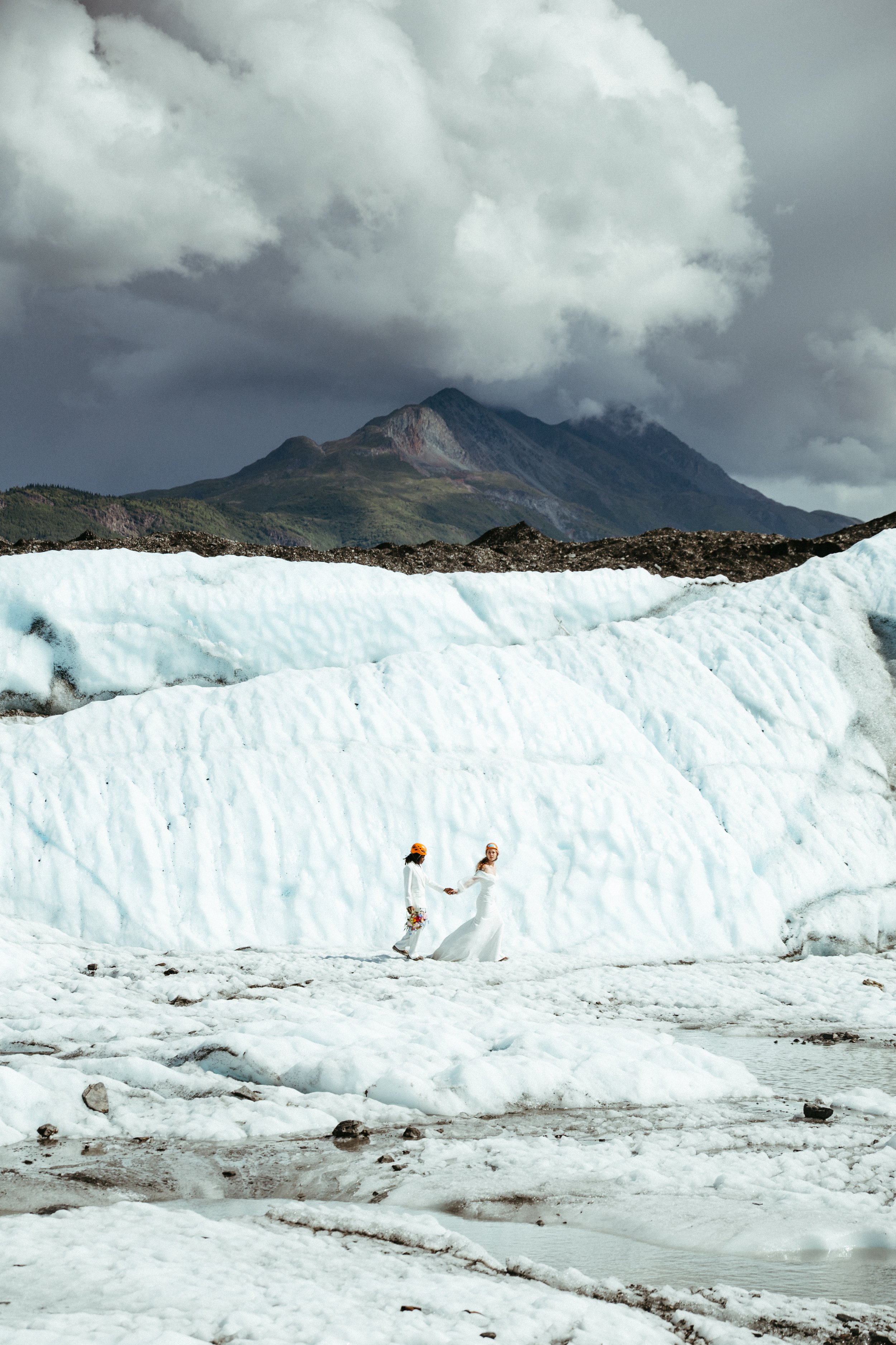 A couple dressed in wedding attire holding hands on a glacier, with mountains and dark cloudy skies in the background.