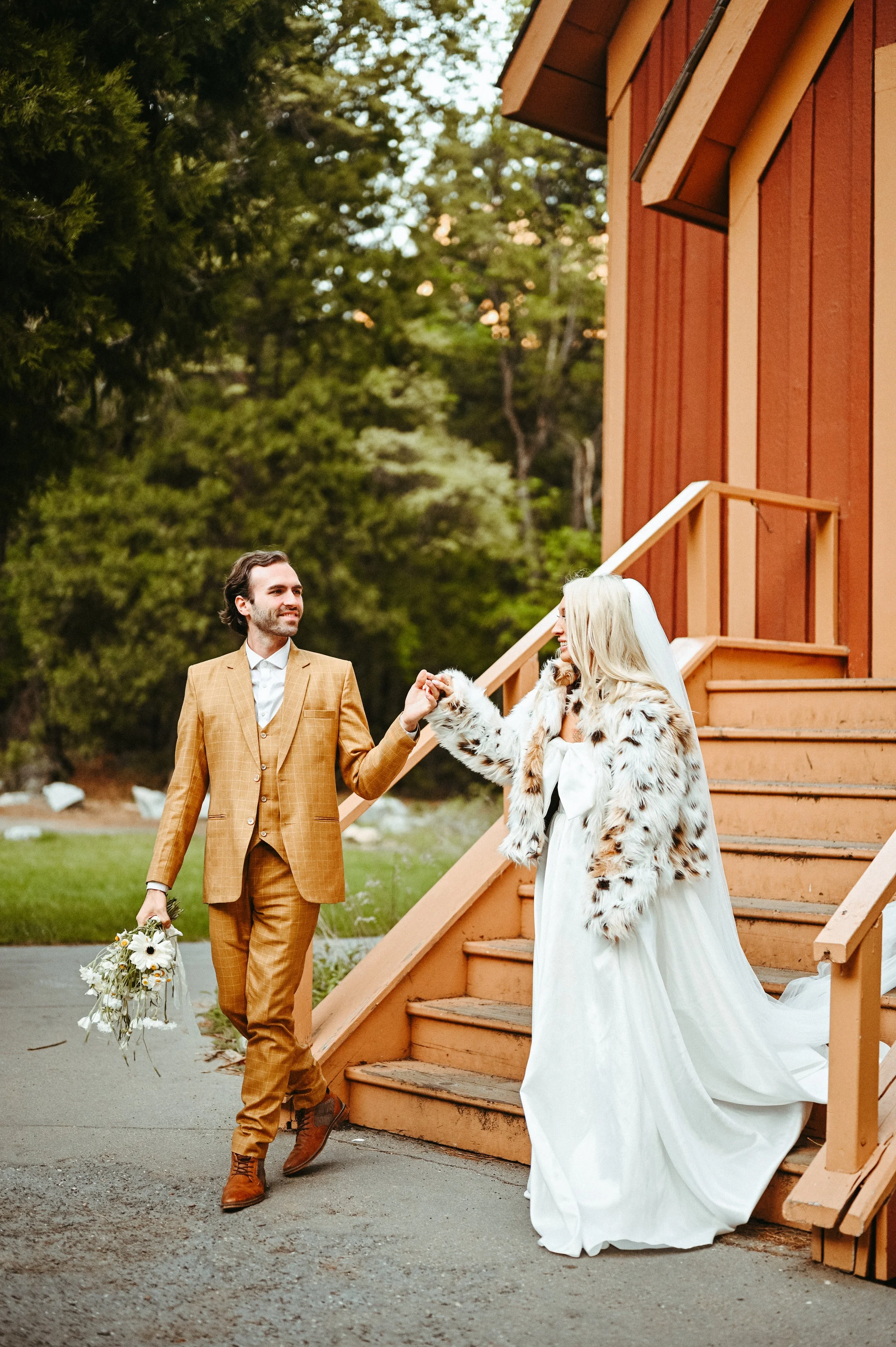 A bride and groom sharing a dance outside at sunset, near a wooden staircase and red barn, with trees in the background.
