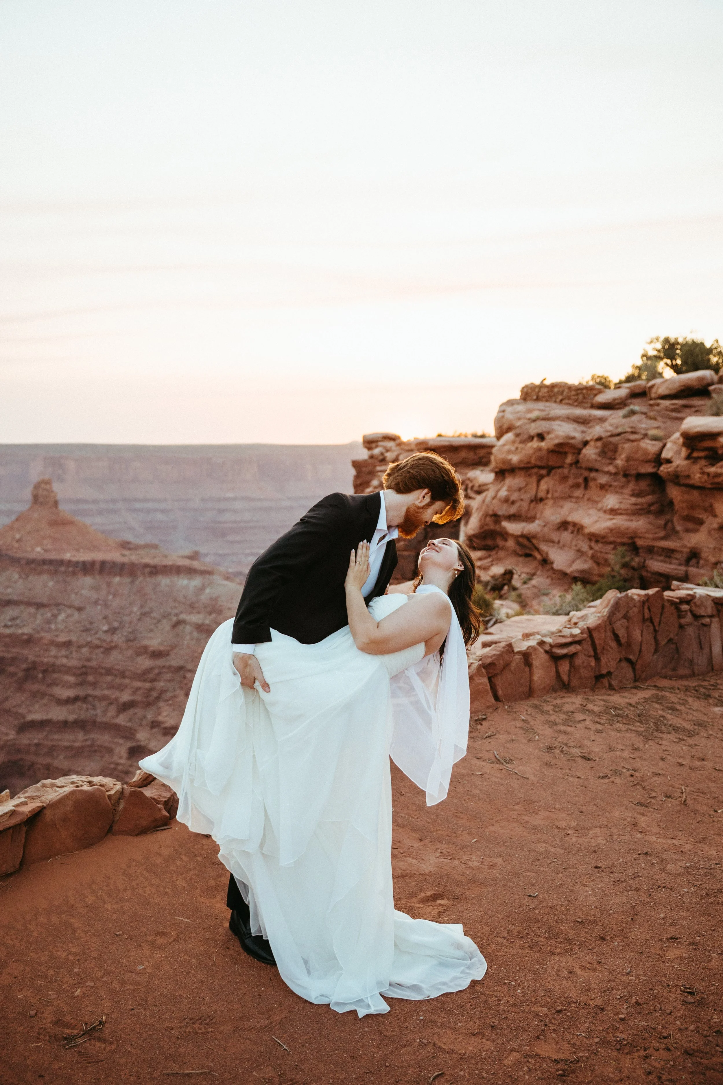 A couple dressed in wedding attire standing on a red rocky landscape at sunset, with someone dips their partner, both smiling and looking at each other.