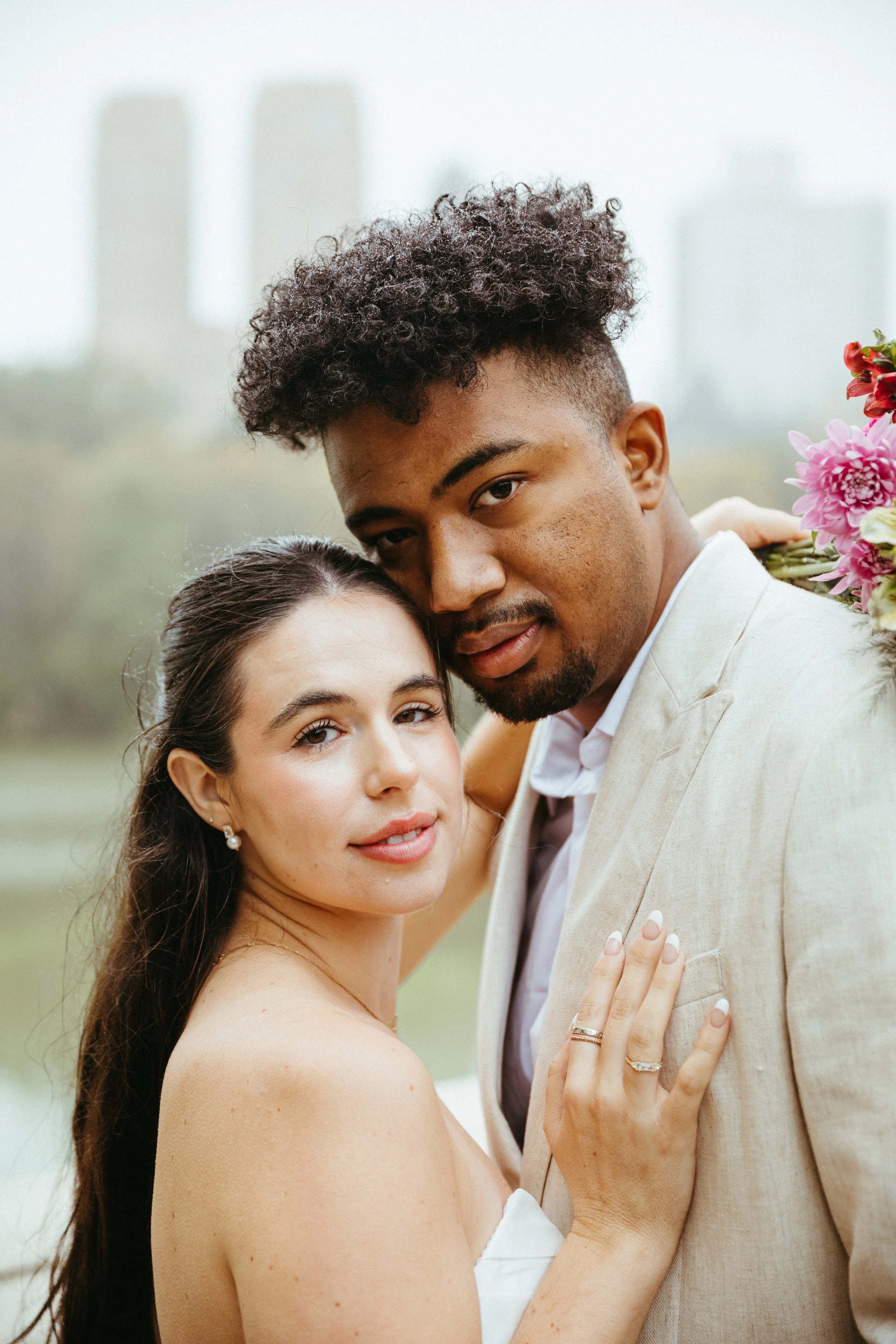 A couple, dressed formally, embracing outdoors with city buildings in the background, holding a bouquet of flowers.