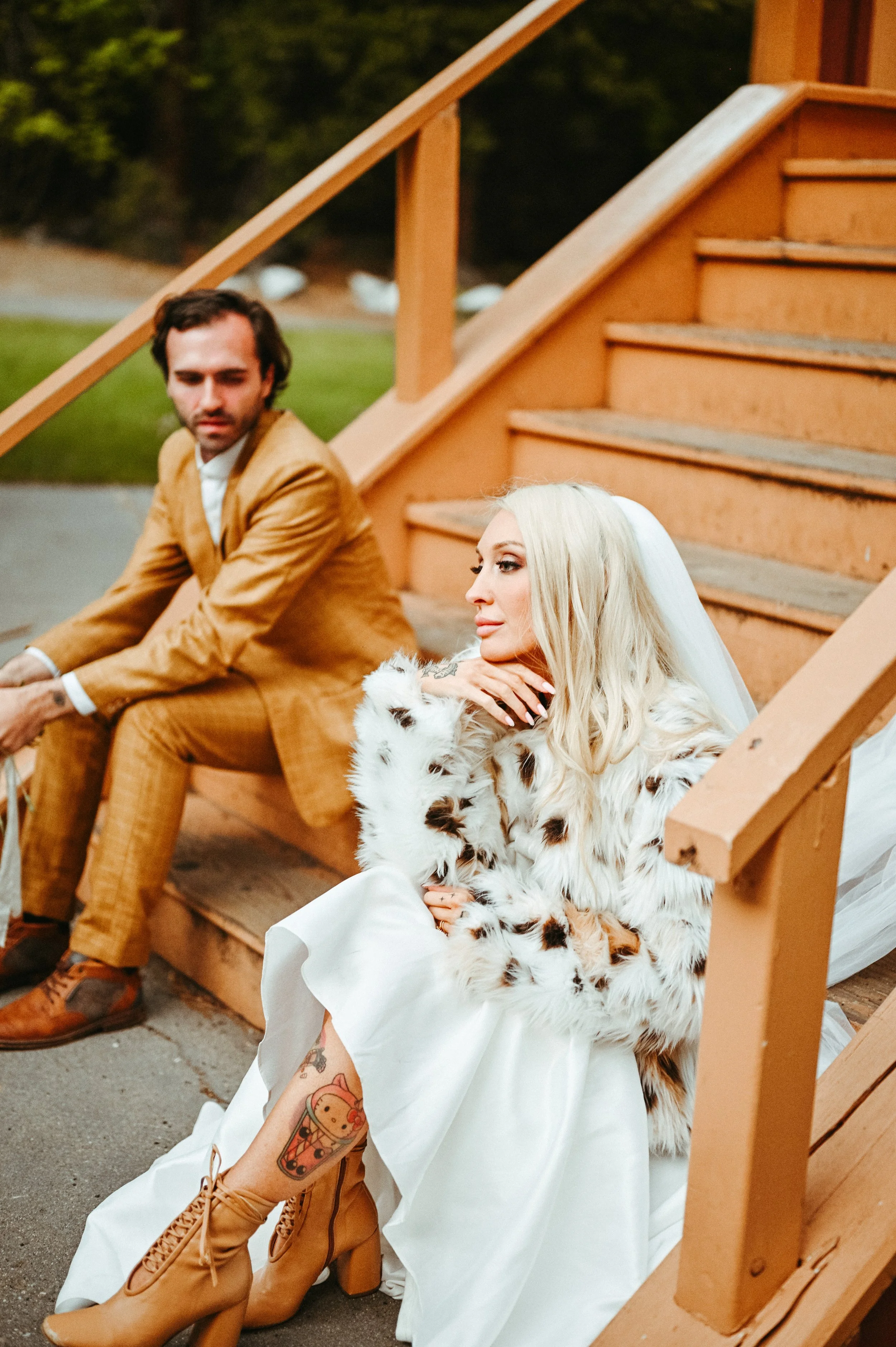 A woman with long blonde hair, tattoos, and wearing a white dress with a fur jacket and beige lace-up boots sits on stairs, with a man in a brown suit sitting nearby.