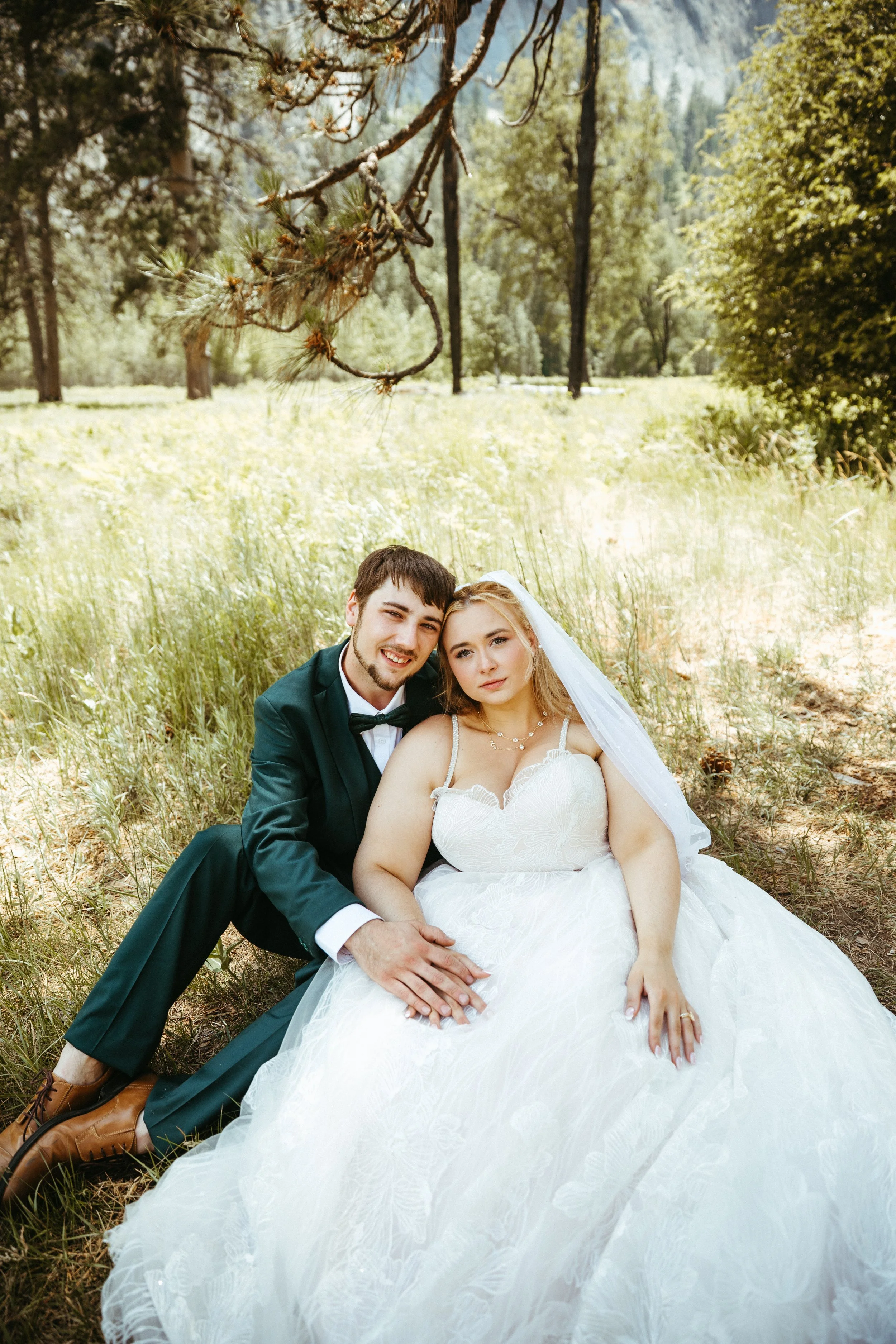 A man and a woman in wedding attire sitting on the grass in a forested area, with tall trees and sunlight in the background.
