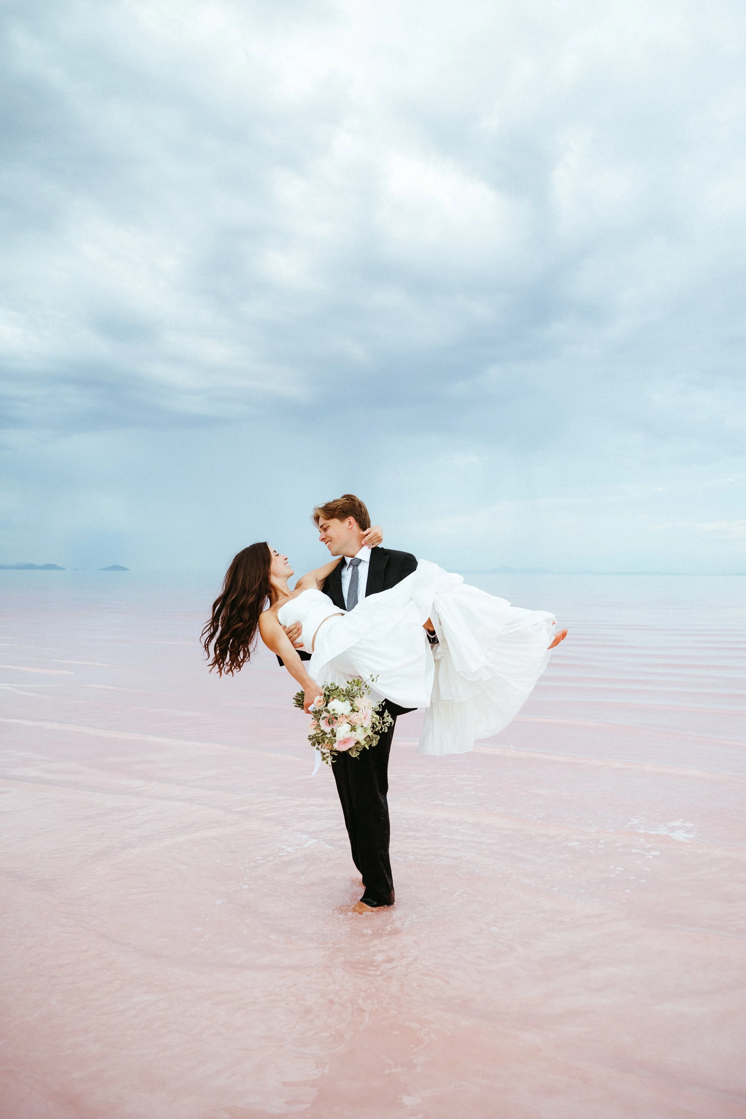 A couple in wedding attire on a beach, with the groom carrying the bride while holding a bouquet of flowers, under a cloudy sky.
