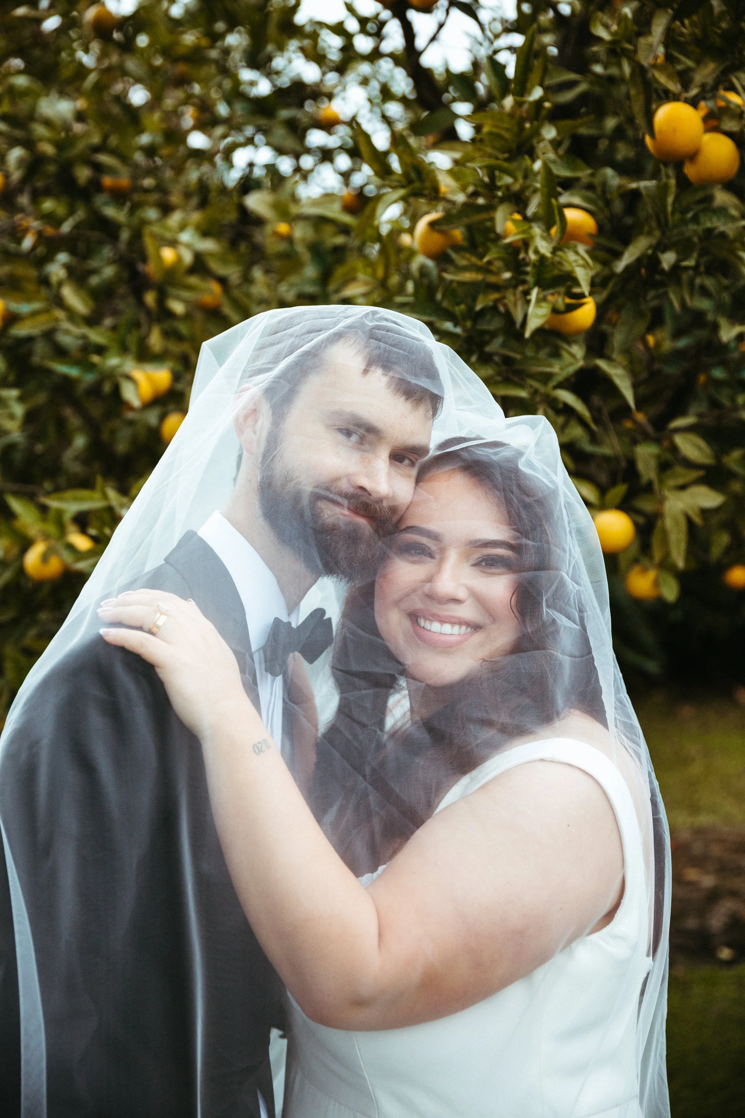A smiling bride and groom embracing behind a wedding veil outdoors near a tree with yellow fruit.