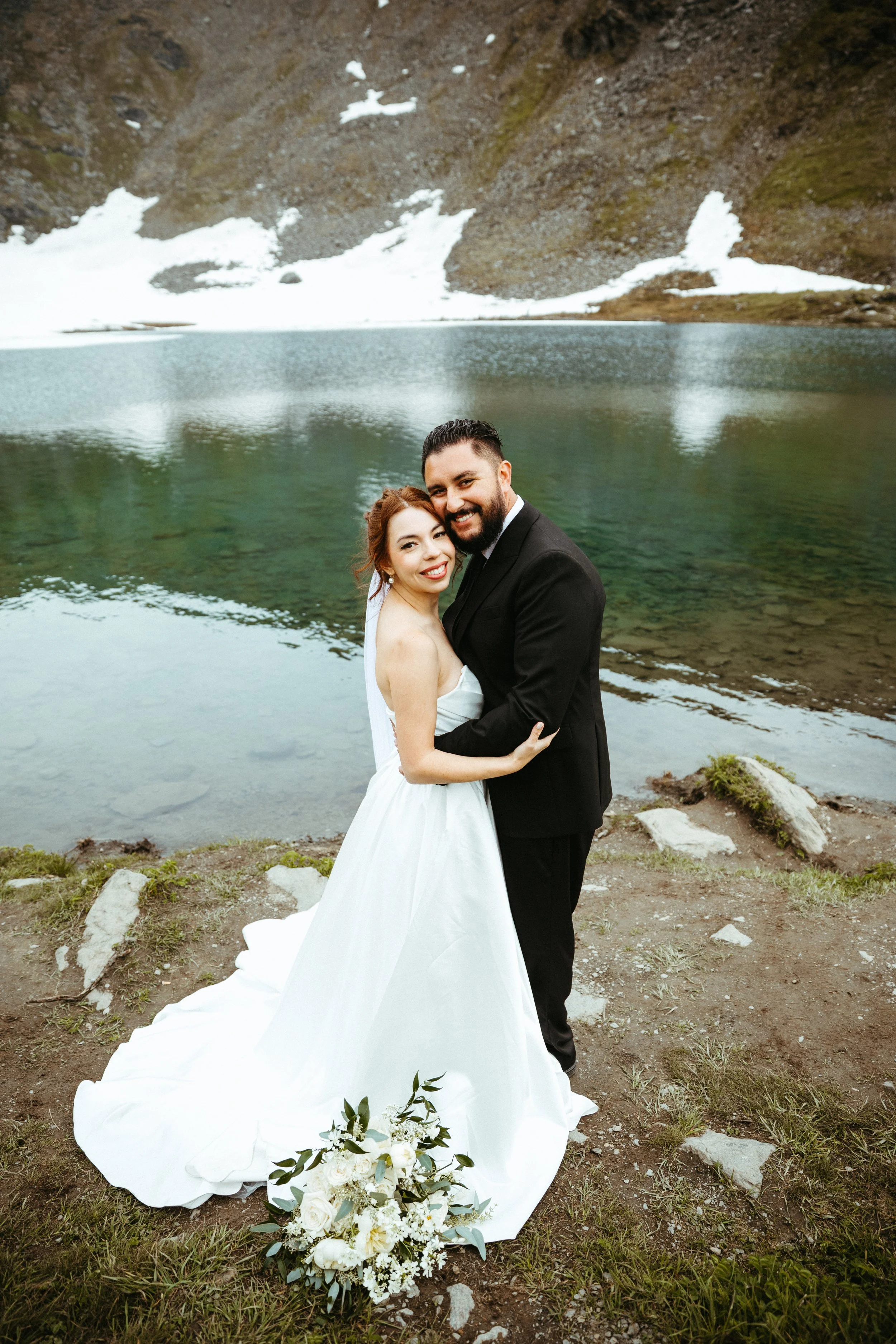 Bride and groom embrace near a mountain lake with snow patches and rocky slopes in the background.