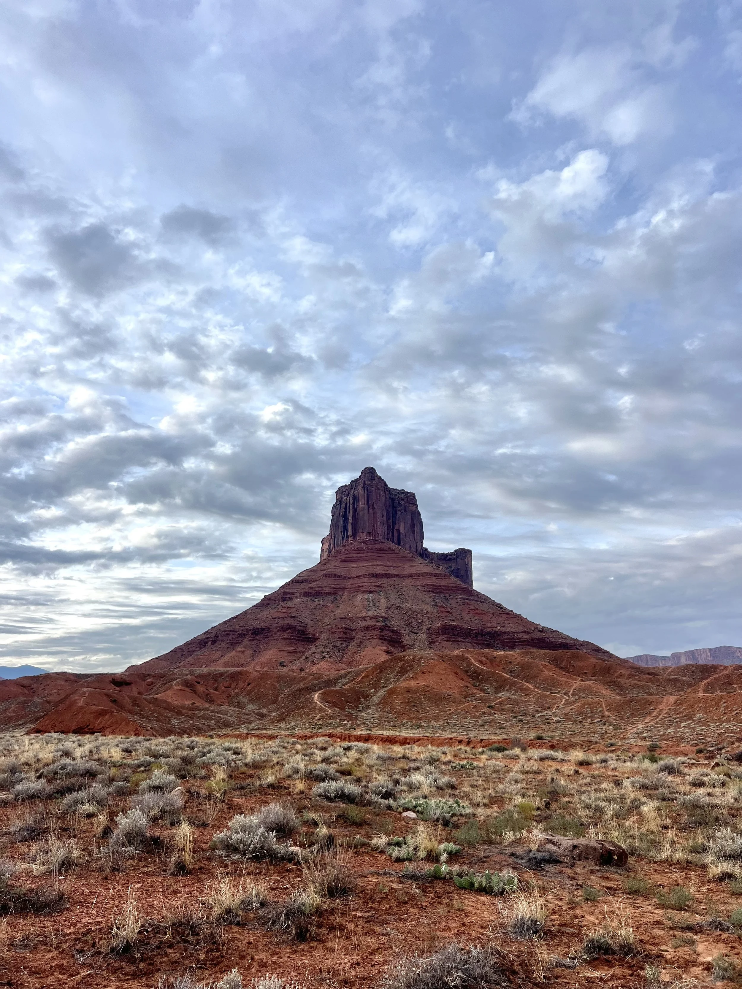 A large, flat-topped red sandstone formation in a desert landscape with sparse vegetation and a cloudy sky.