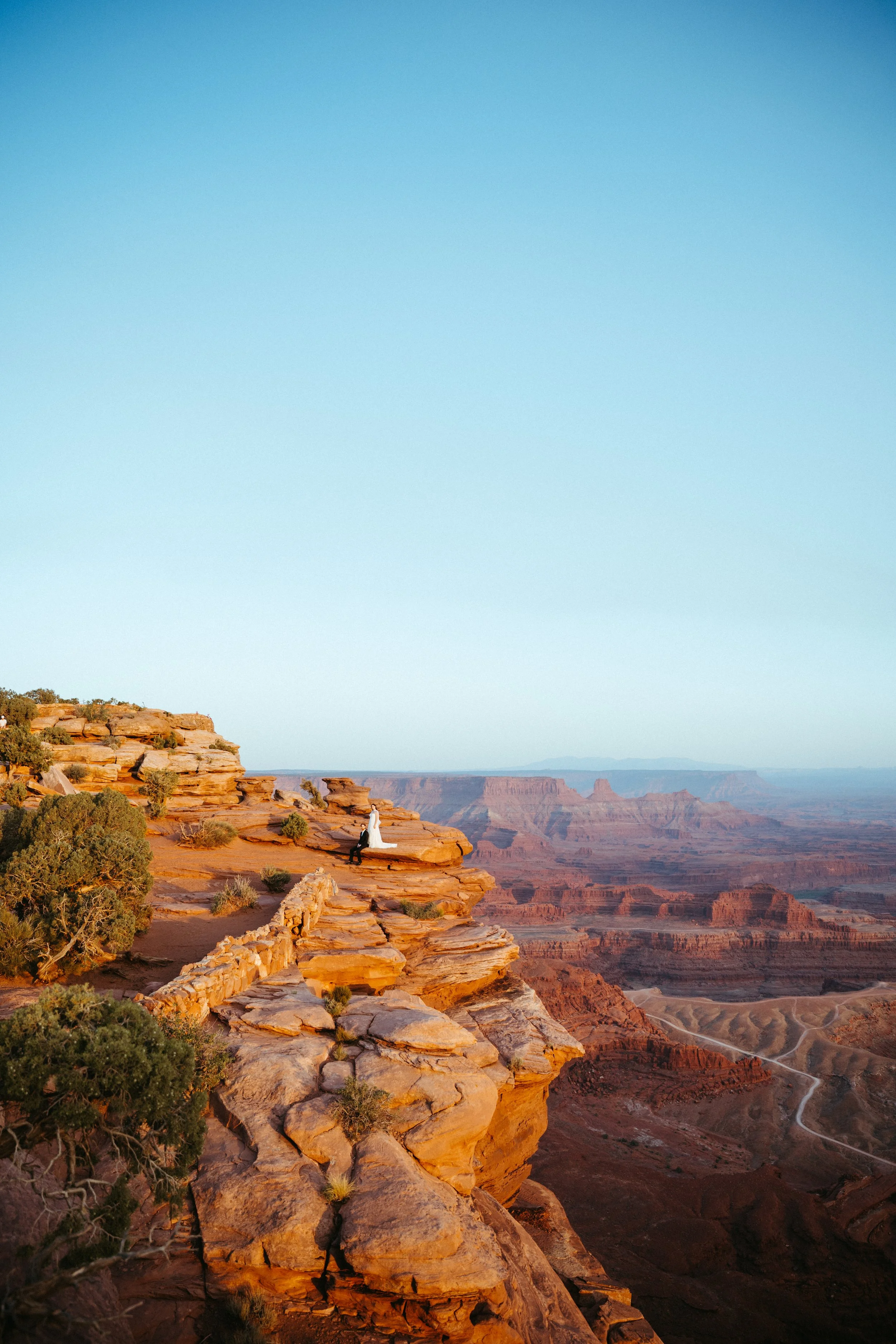A couple in wedding attire sitting on a rocky ledge overlooking the Grand Canyon at sunset.