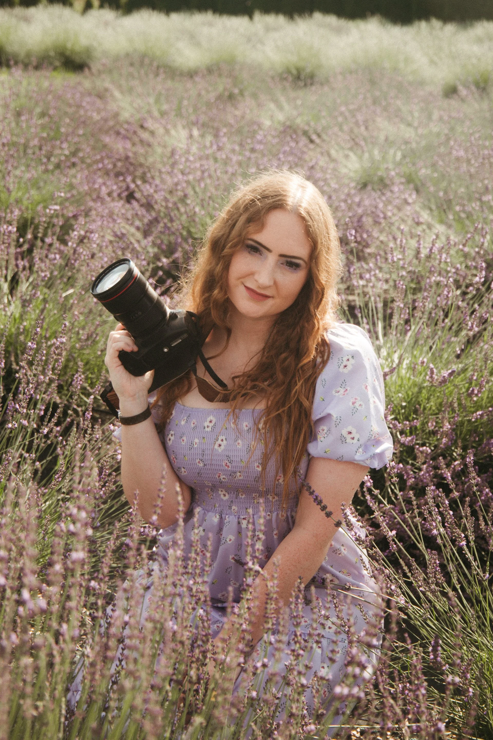 A young woman with long, curly red hair, wearing a purple floral dress, standing in a field of blooming lavender flowers, holding a professional camera.