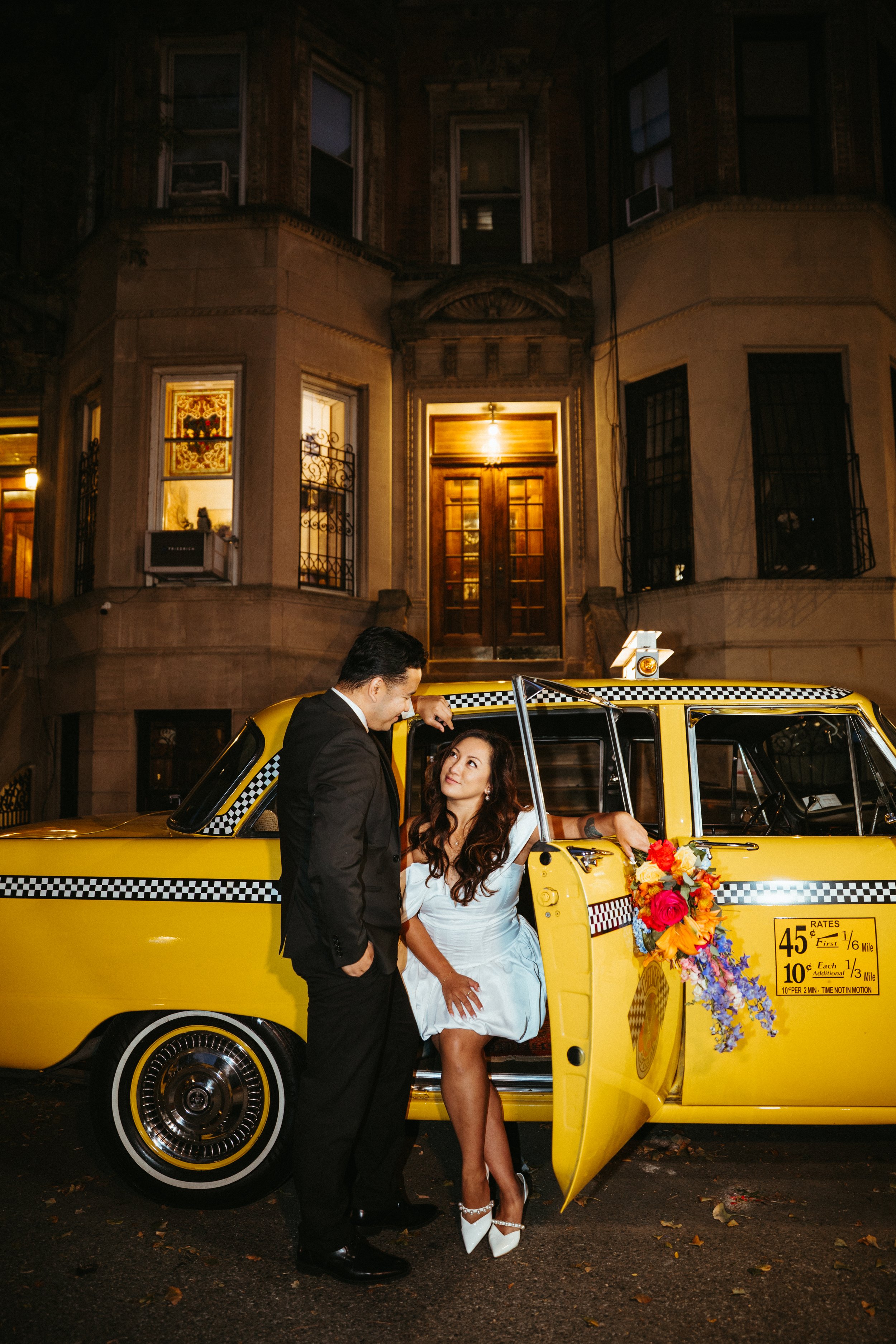 A couple dressed in formal attire stands outside a yellow taxi cab with a floral arrangement on the door. The woman is sitting inside the cab, looking up at the man, while the man leans against the cab's roof, engaging with her. The background features a brownstone building at night.