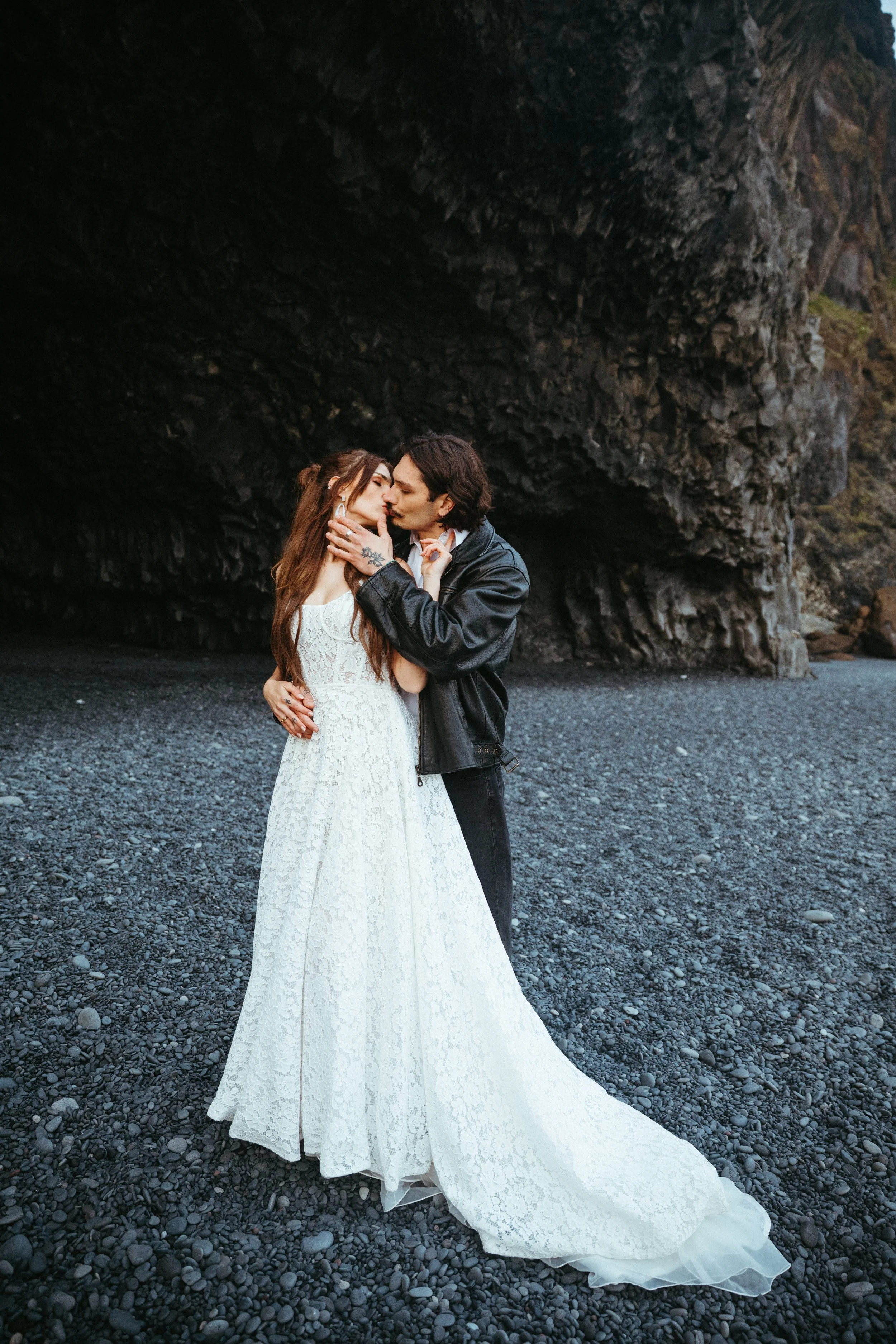 A couple sharing a kiss on a rocky beach, with the woman in a white wedding dress and the man in a black leather jacket.